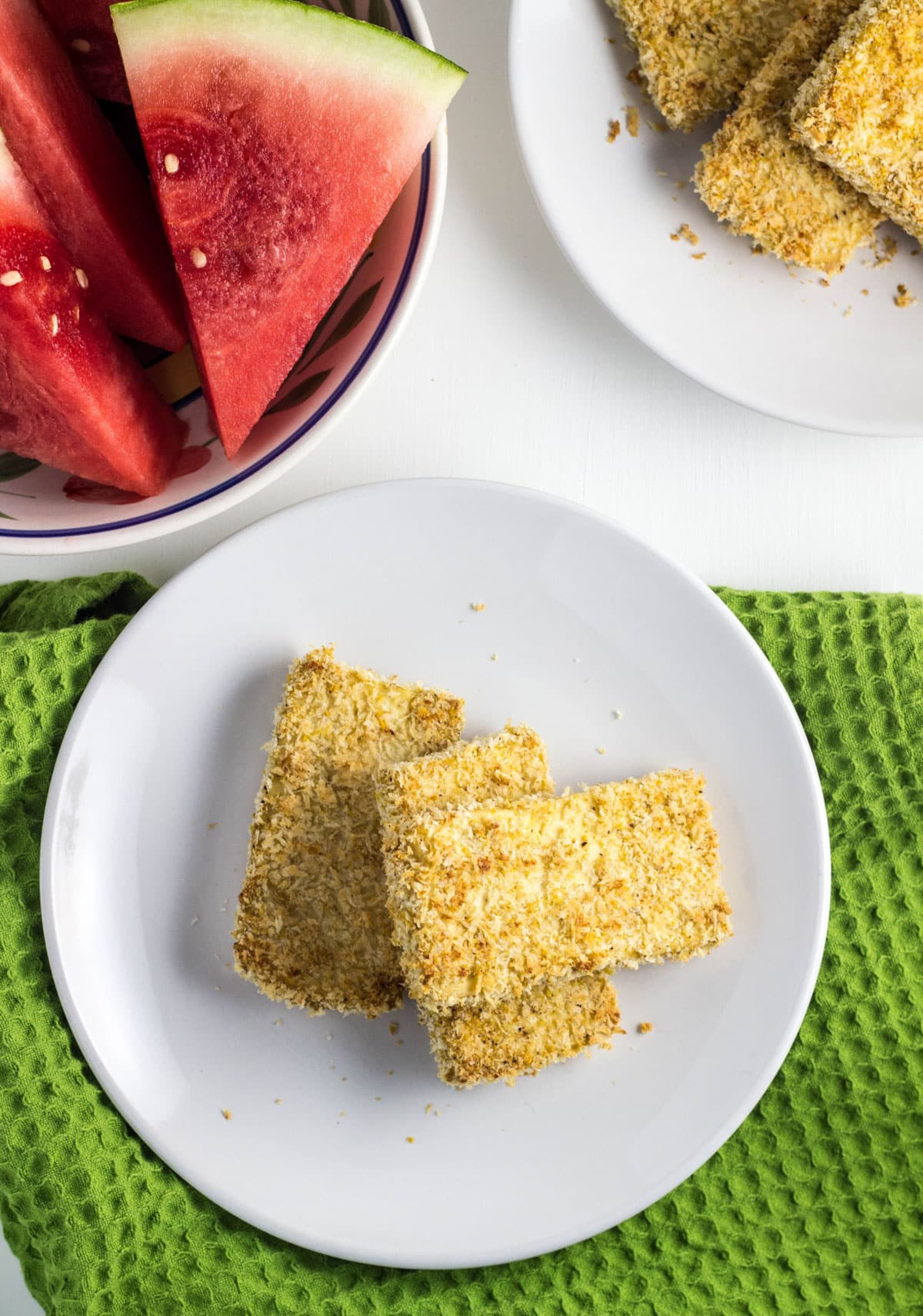 Overhead view of a white plate with three Breaded Tofu Sticks. There is a bowl of watermelon partially in view.