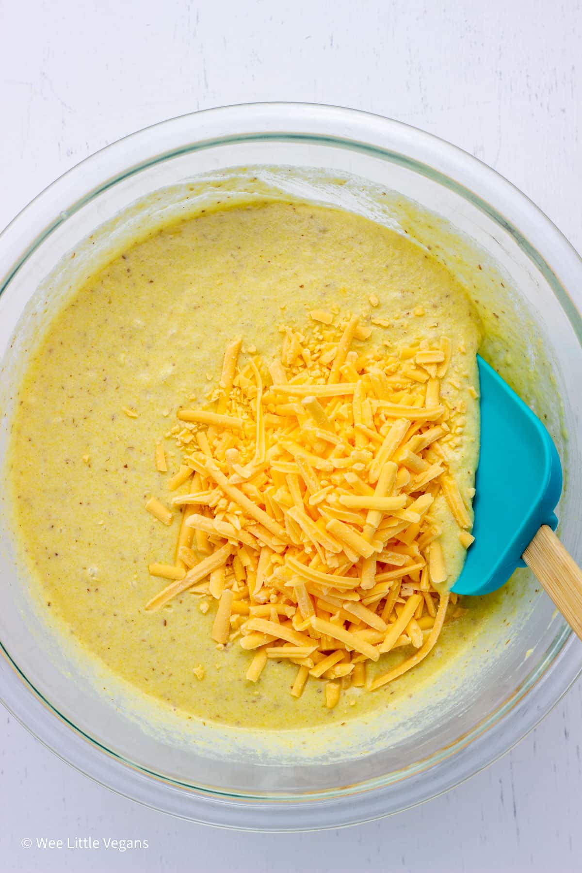 Overhead shot of Cornbread Waffle batter in a bowl. There is non dairy cheese on top of the batter with a rubber spatula to the side.
