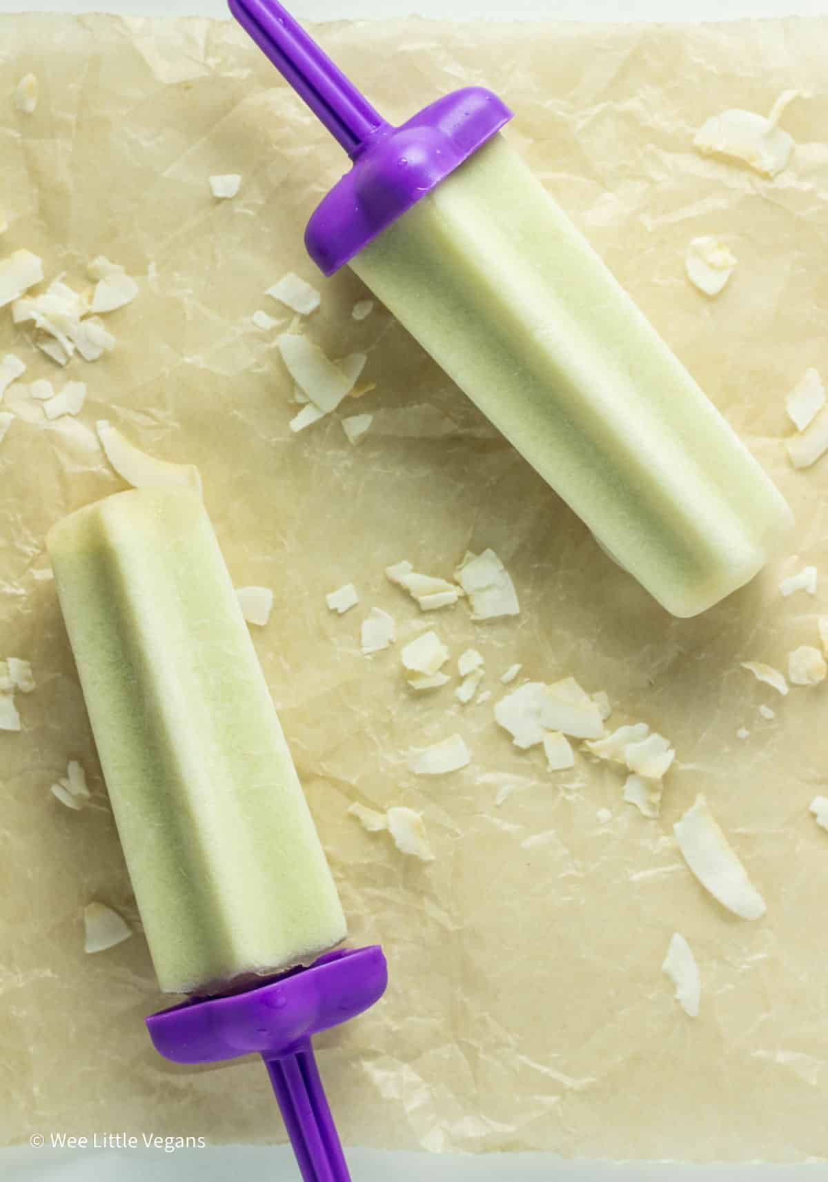 Overhead view of two Honeydew Melon Popsicles laying on parchment paper with coconut flakes scattered around.