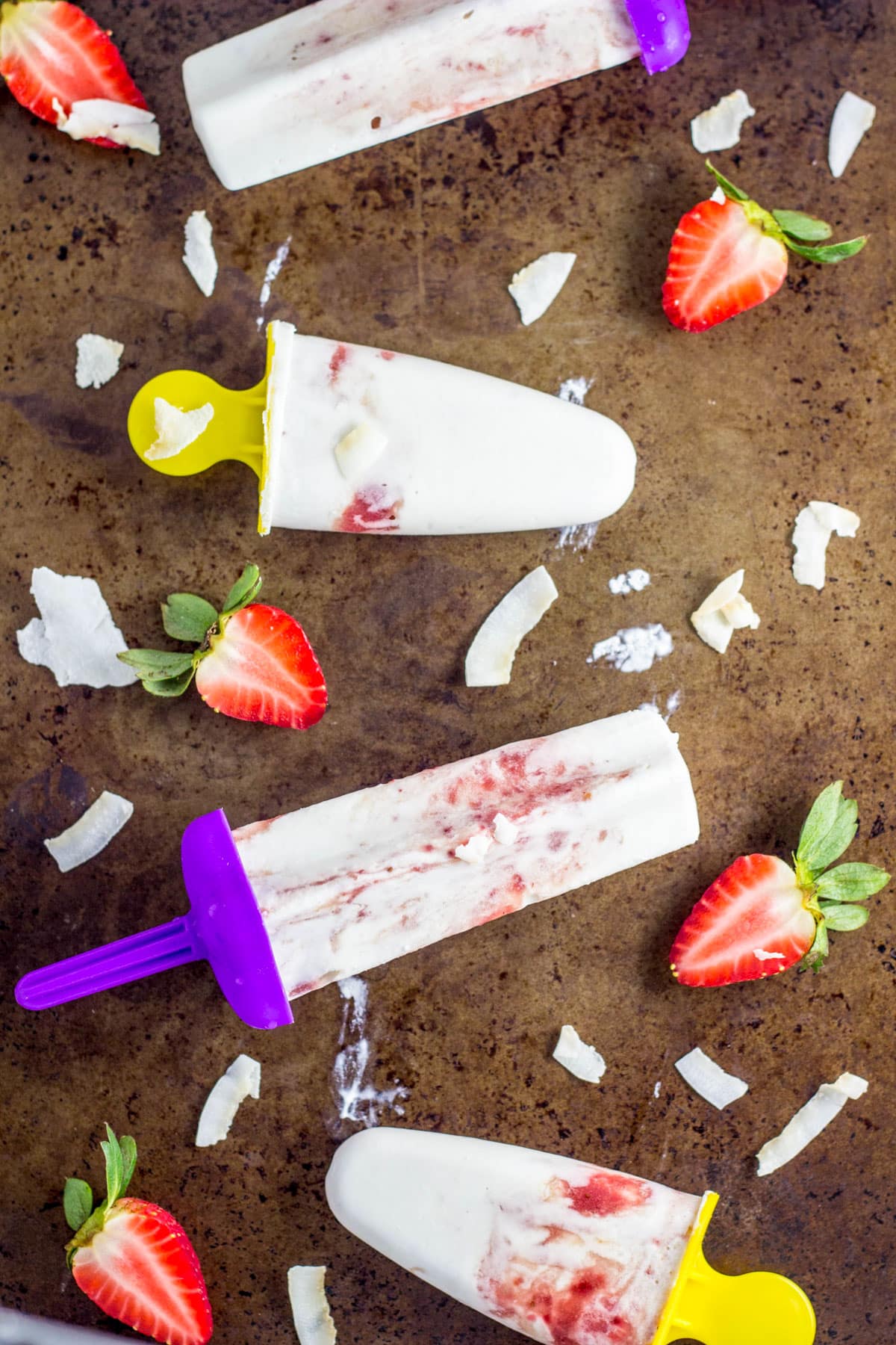 Overhead shot of Strawberry Coconut Cream Pops on a distressed baking sheet. There are strawberry halves and coconut chips around the pops.