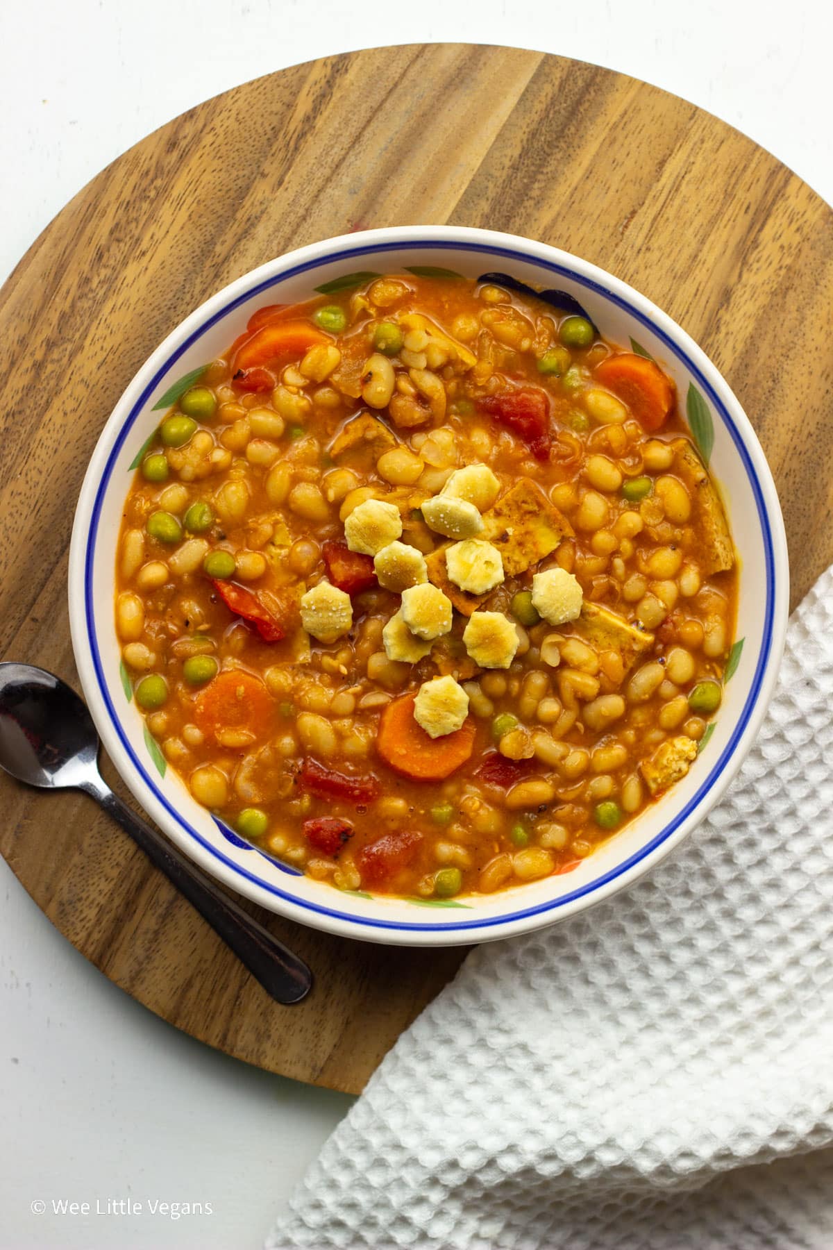 Overhead view of a bowl of Vegan Navy Bean Soup. The bowl sits on a round wooden board and there are small crackers on top of the soup.