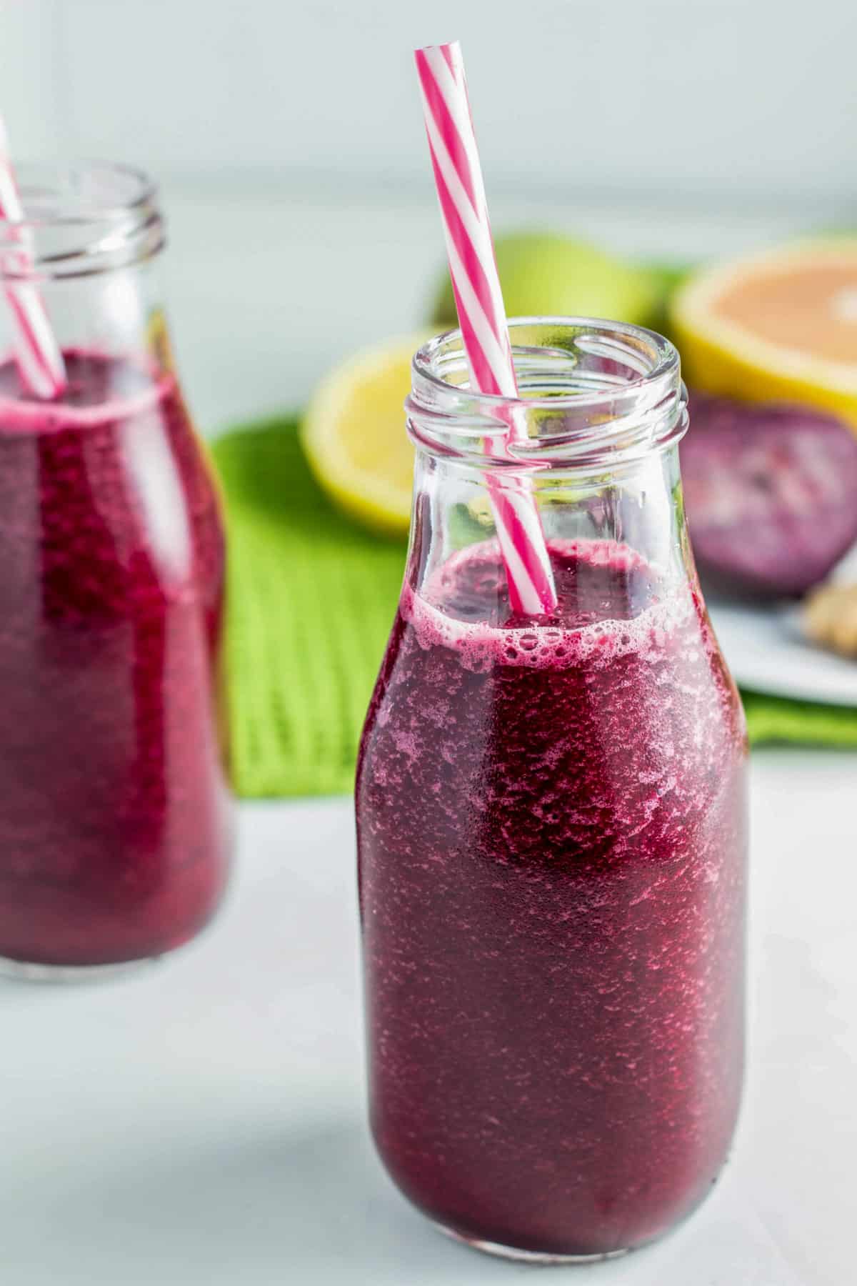 Side view of a bottle of Beet Citrus smoothie with pink and white striped straw. There is another bottle and fruit in the background.