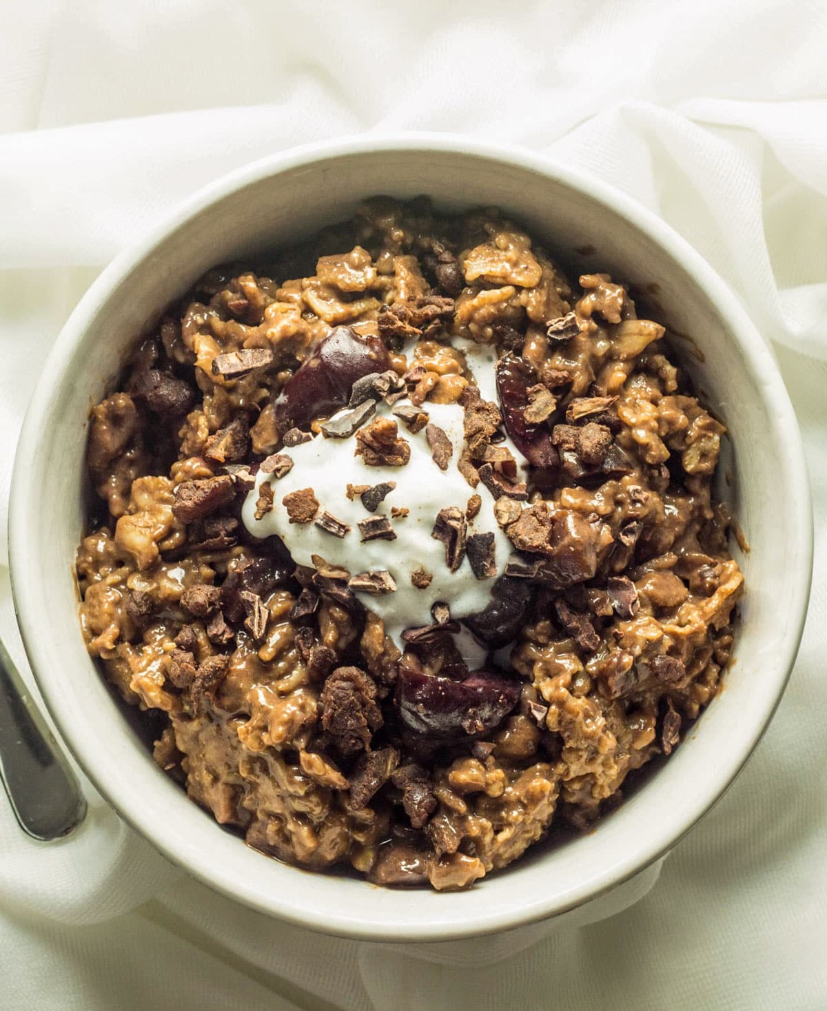 Overhead shot of Black Forest Cake Oatmeal in a white bowl. There is coconut whipped cream on top with cacao nibs sprinkled over.