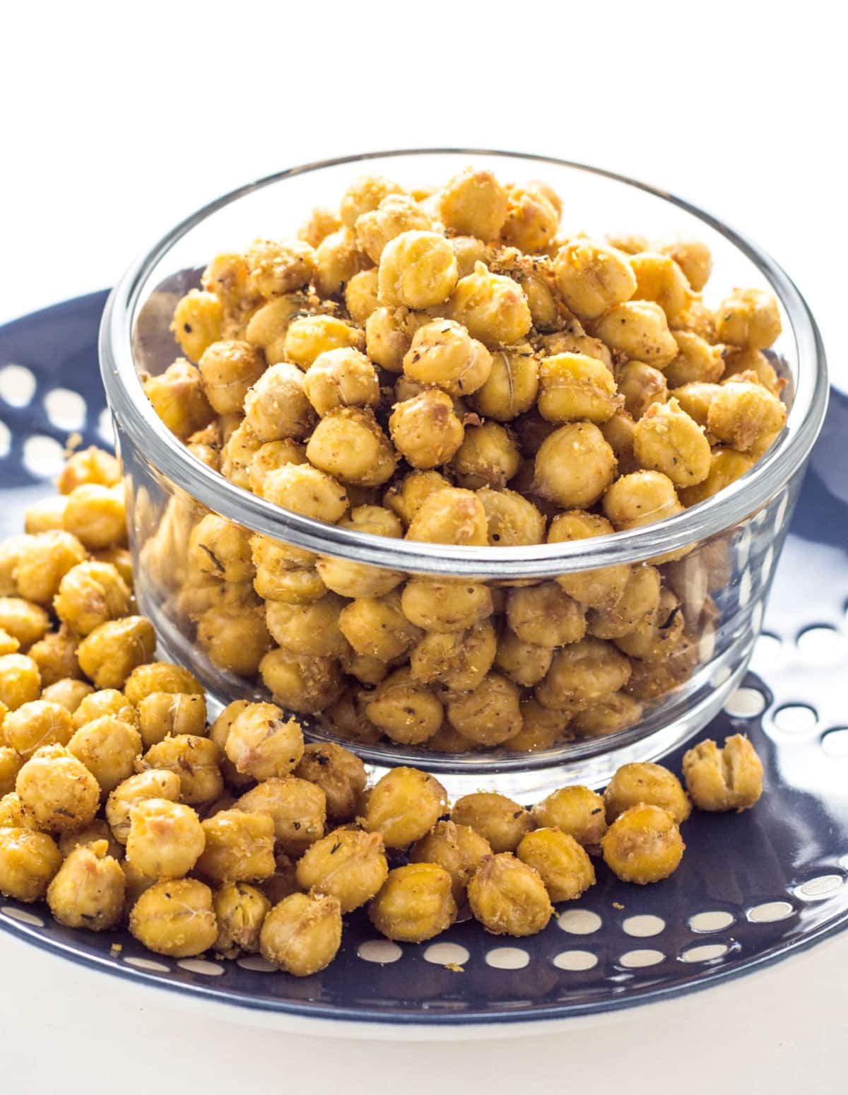 A bowl of Cheezy Herb Roasted Chickpeas sitting on a blue dotted plate. There are more chickpeas on the plate.