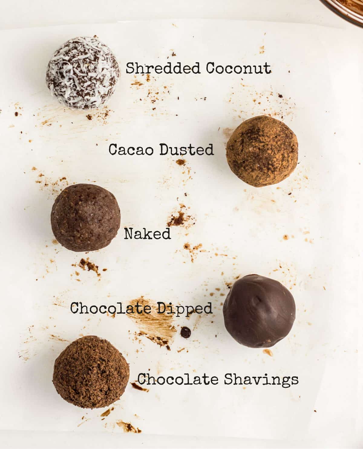 Chocolate Coconut Butter Truffles on parchment paper. There is text next to each truffle describing the coating on the exterior coatings including shredded coconut, cacao dusted, naked, chocolate dipped and chocolate shavings.