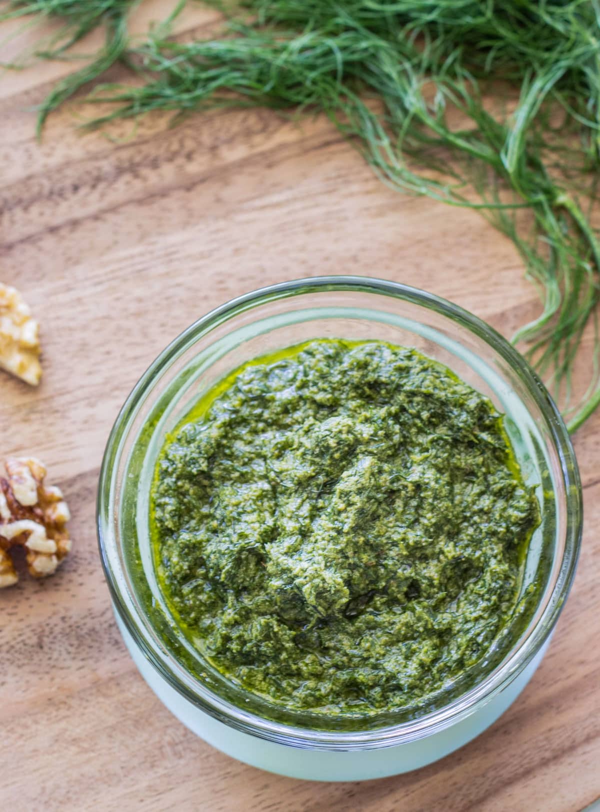 Overhead view of Fennel Walnut Pesto in a small bowl. The bowl is sitting on a wooden board and there walnuts and fennel fronds partially visible in the frame.