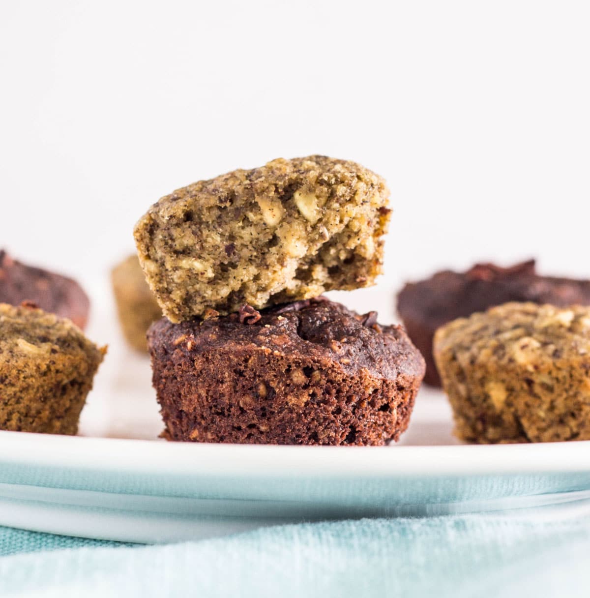 Close up of Gluten Free Oil Free Mini Banana Muffins stacked. The bottom muffin is chocolate. The top muffin is half of a banana bread muffin showing the interior.