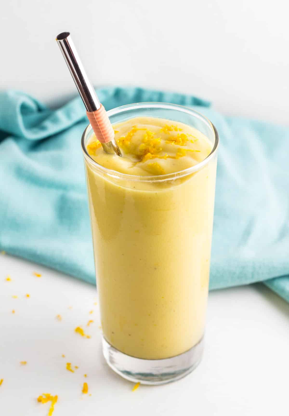 A tall glass Of Orange Mango Smoothie with a straw. There is a light blue cloth in the background.
