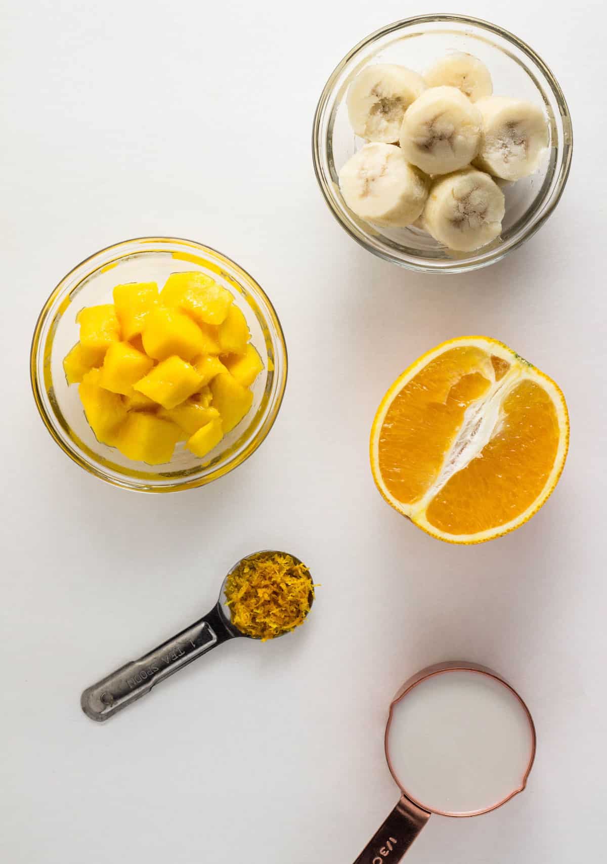 Overhead shot of ingredients to make an Orange Mango Smoothie. Frozen banana, diced mango orange juice and zest and non dairy milk.