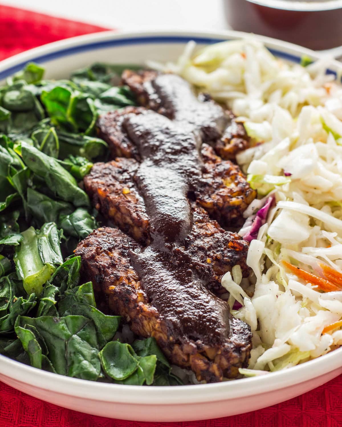 Close up of Vegan Barbecue Tempeh Bowl. There are sautéed greens to the left, barbecue tempeh in the middle and coleslaw to the right of the image.