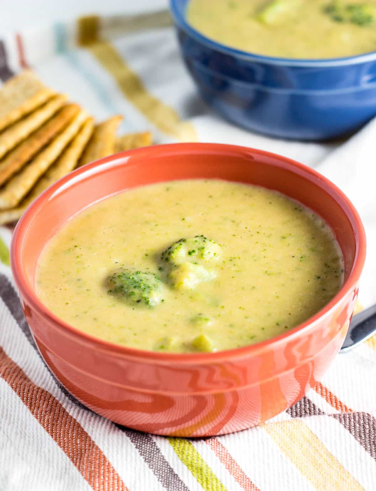 A small orange bowl of Vegan Broccoli Cheese Soup in the foreground. In the background there is a a second blue bowl of soup.