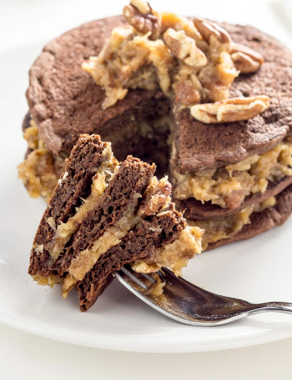 Close up of a bite of German Chocolate Pancakes on a fork resting on a plate. The rest of the stack is behind the fork on the plate.