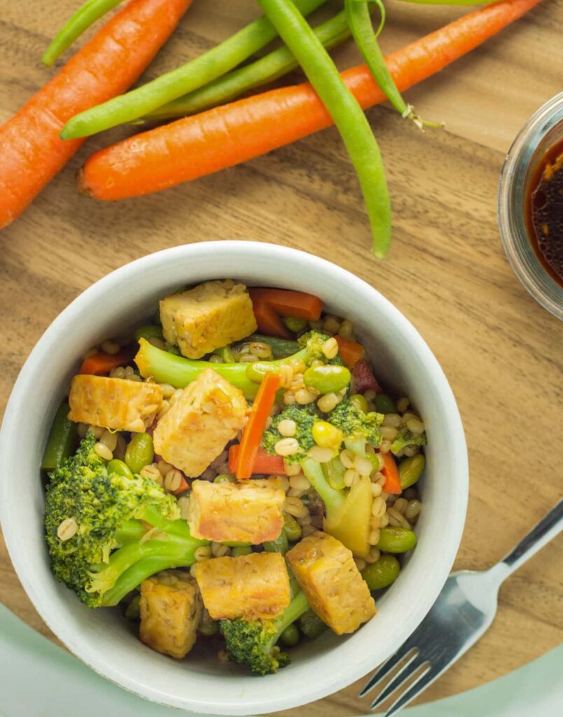 Overhead view of Vegan Teriyaki Tempeh Bowl. There are fresh carrots, green beans and a small bowl of sauce partially visible to the sides of frame.