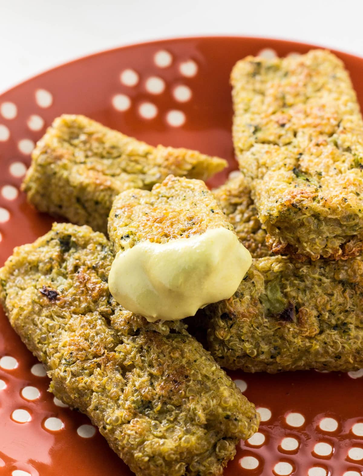 Close up view of a Broccoli Quinoa Stick dipped in cashew cheese sauce. There stick rests on a stack of other sticks on an orange and white plate.