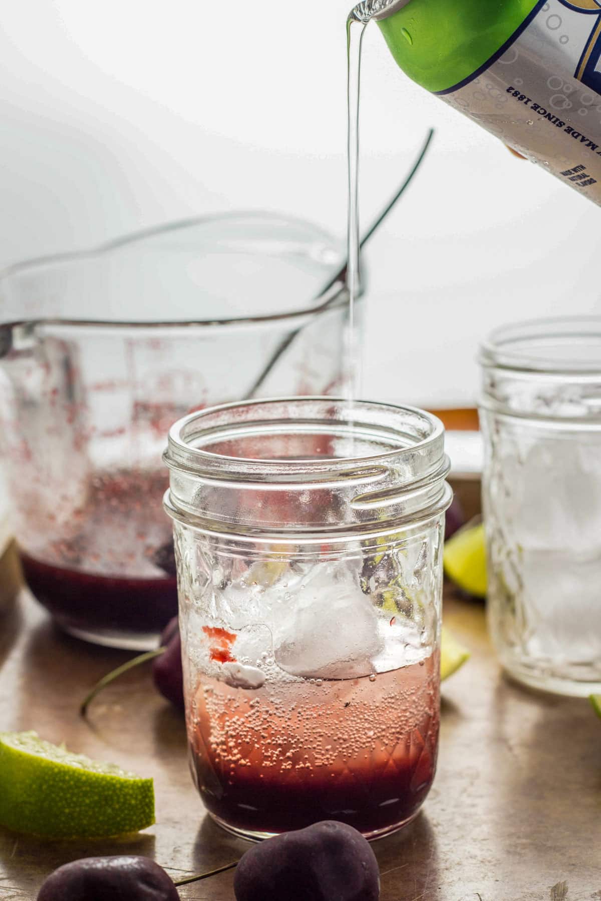 A can of seltzer pouring into a glass with cherry syrup to make a Cherry Lime Spritzer.