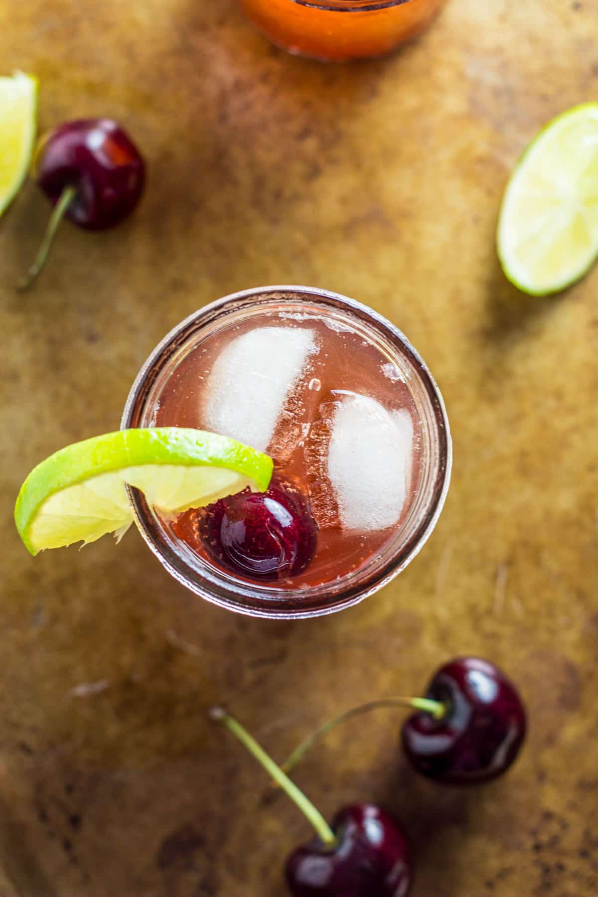 Overhead view of a glass of Cherry Lime Spritzer. There are ice cubes and a cherry in the glass and a wedge of lime on the edge of the glass.