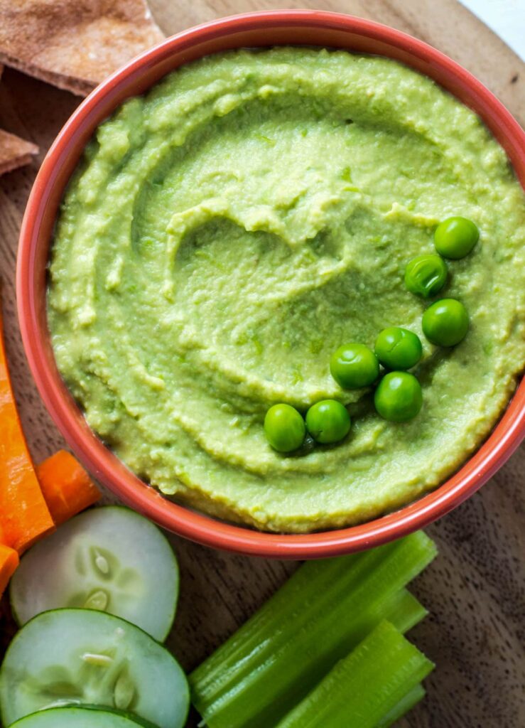 Overhead view of a small orange bowl of Green Pea Hummus. Cucumber slices, celery and carrot sticks are partially visible in the bottom right corner.