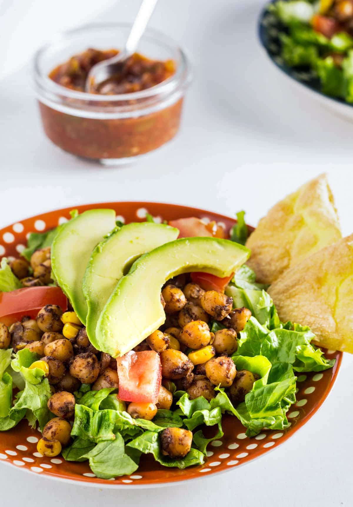 Close up side view of Vegan Chickpea Taco Salad on an orange and white plate. The salad consists of a bed of romaine lettuce topped with seasoned chickpeas, corn kernels, diced tomatoes, avocado and tortilla chips.