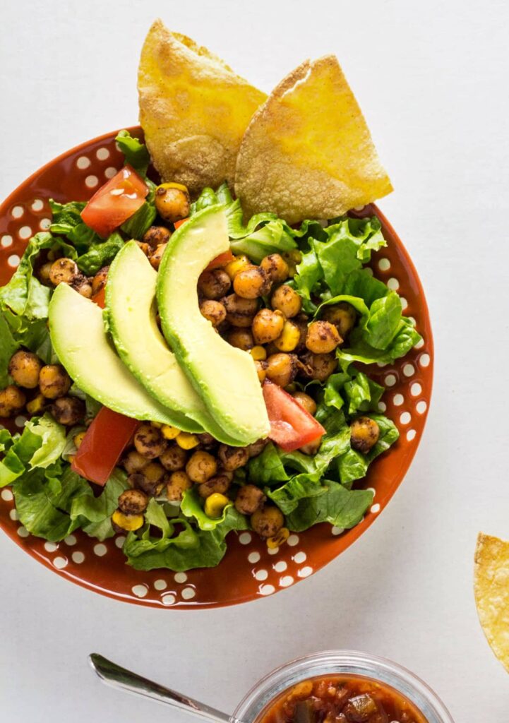 Overhead view of Vegan Chickpea Taco Salad on an orange and white plate. The salad consists of romaine lettuce topped with seasoned chickpeas, corn kernels, diced tomatoes and avocado with tortilla chips on the side of the dish.