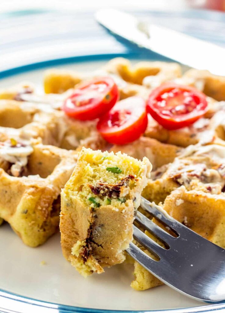 Close up of a fork with a bite of a Vegan Savory Waffle. The fork is resting on the edge of a plate with the remaining waffle.