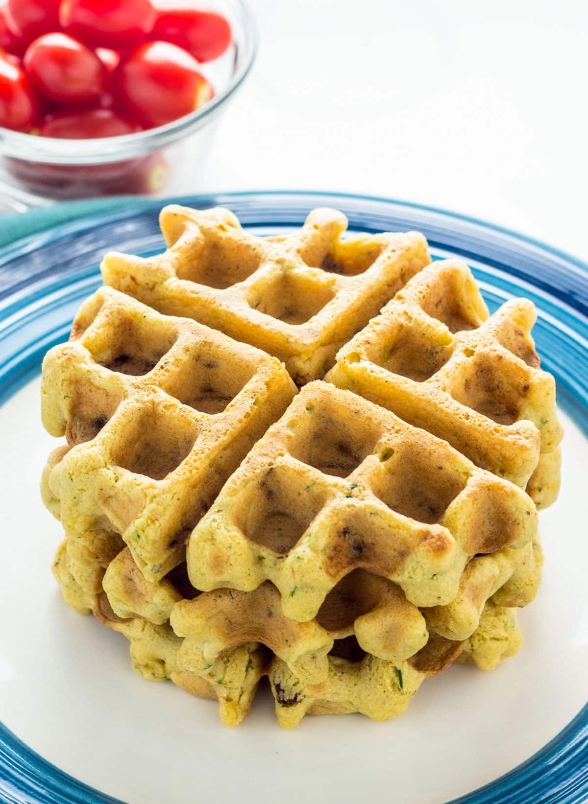 Three Vegan Savory Waffles stacked on a plate. There is a small bowl of grape tomatoes in the background.