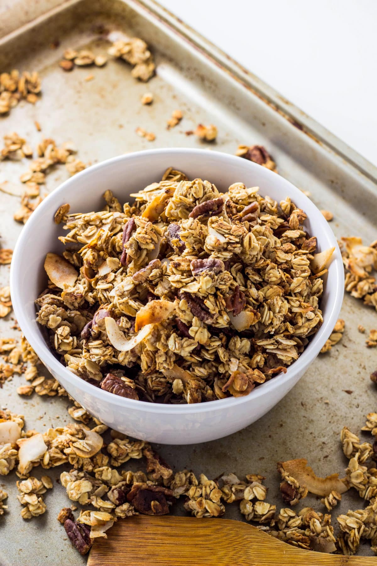 A bowl of Coconut Pecan Banana Granola sitting on a baking sheet with more granola around the bowl.