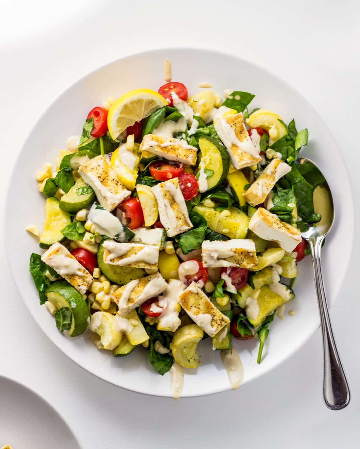 Overhead view of a plate of Grilled Tofu Summer Veggie Salad. Vegetables include zucchini, yellow squash, tomato, corn and spinach. The salad is dressed with a lemon tahini sauce.