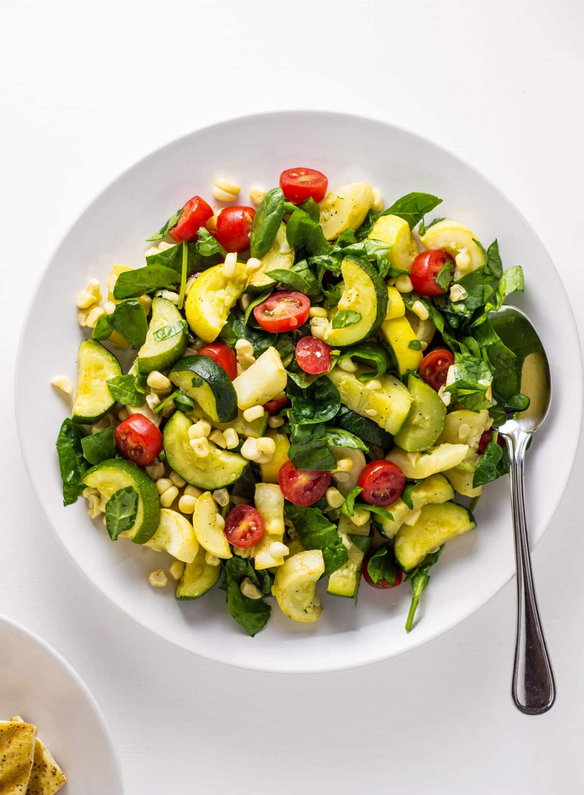 Overhead view of zucchini, yellow squash, tomatoes, corn and chopped spinach tossed together on a white plate. There is a spoon to the side of the plate.