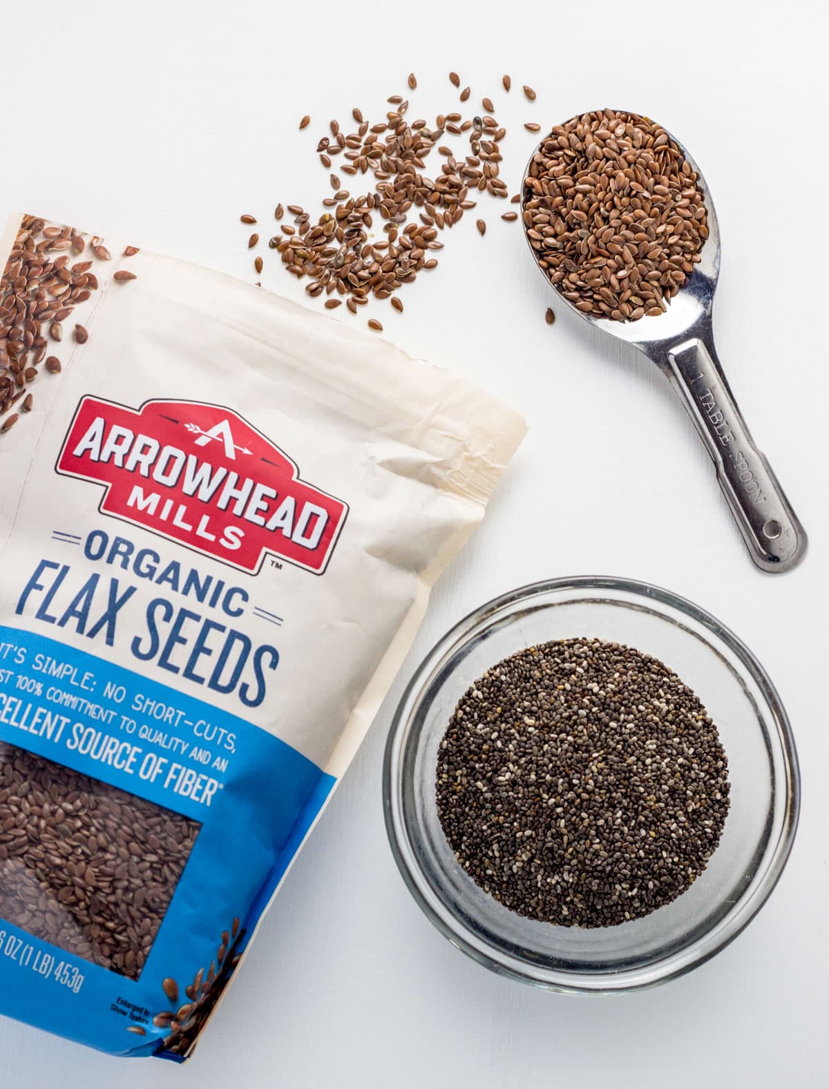 Overhead view of a bag of flax seeds with a spoon of seeds in the upper right. In the lower right corner is a bowl of chia seeds.
