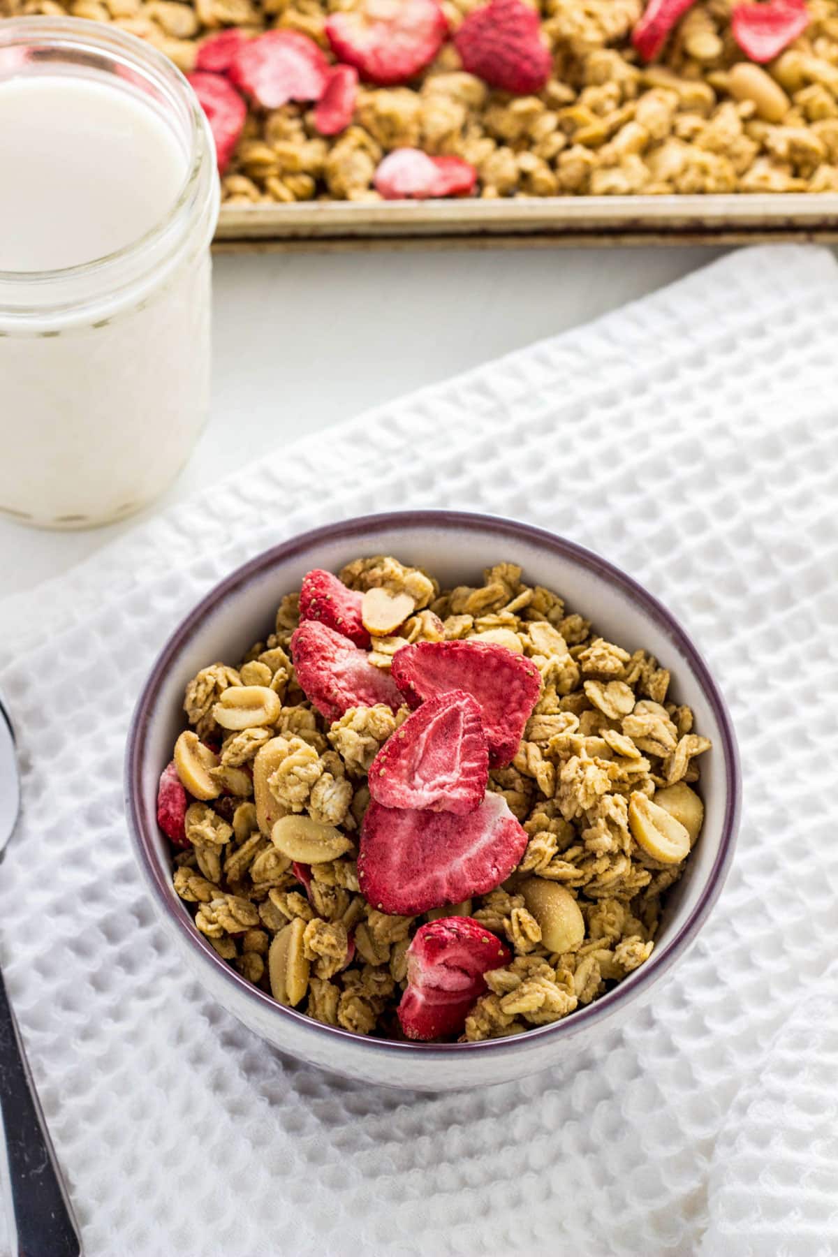Peanut Butter & Jelly Granola in a small bowl. There is a glass of non dairy milk and baking sheet of more granola in the background.