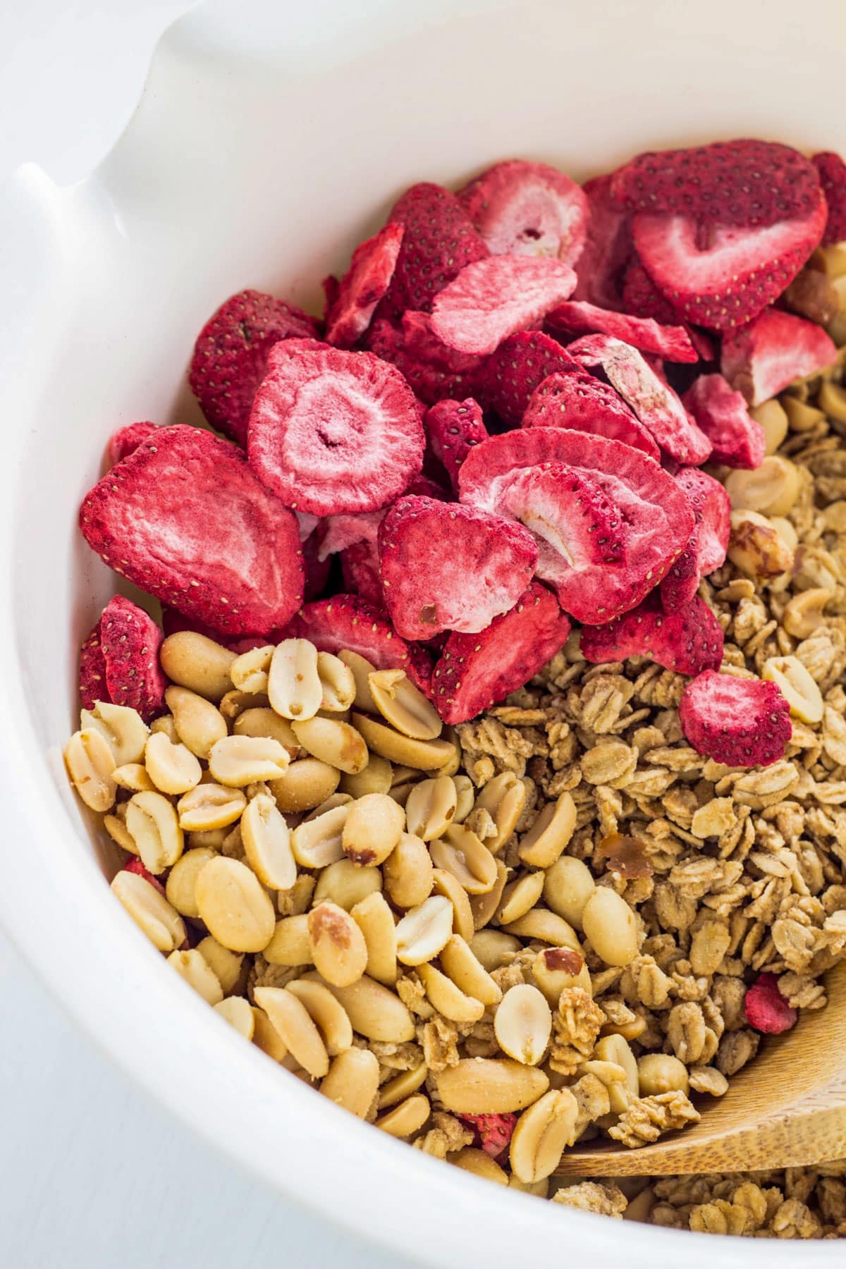 Oats, roasted peanuts and freeze dried strawberries in a large white bowl. The ingredients have not been mixed together. There is a wooden spoon in the bottom right of the image.