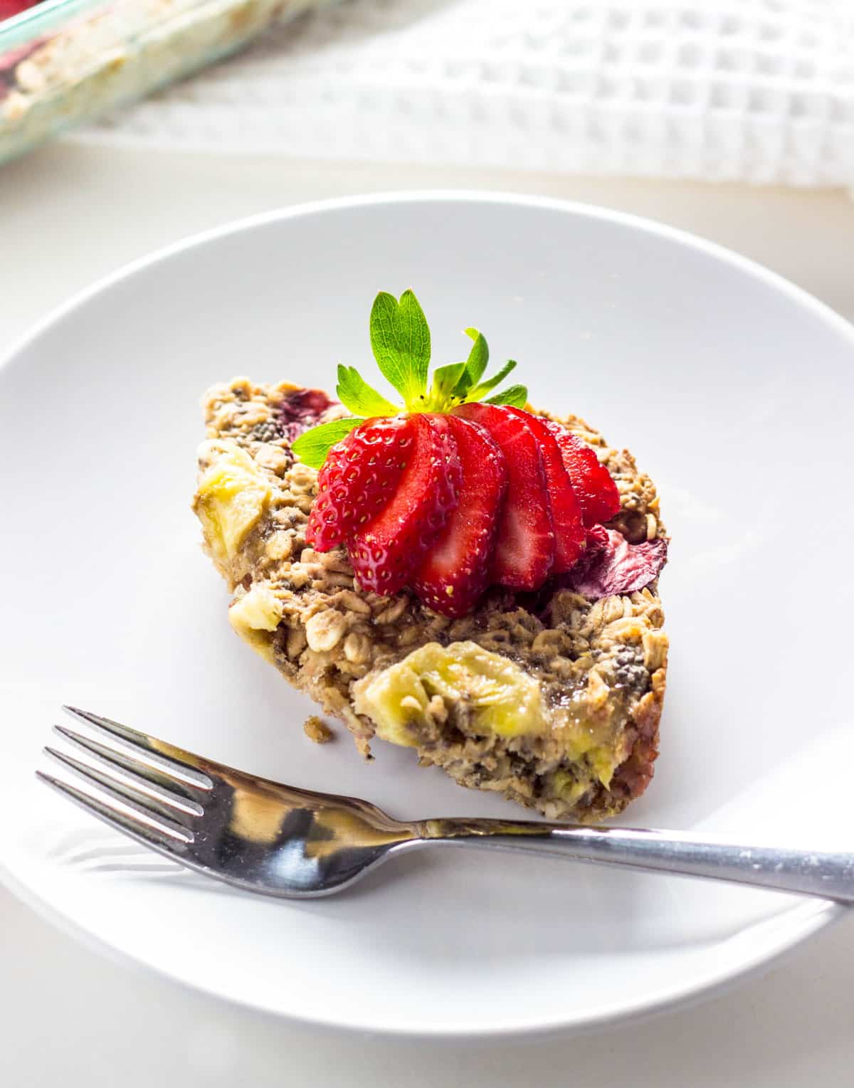 Strawberry Banana Baked Oatmeal on a small white plate. There is a fresh sliced strawberry on top of the oatmeal.