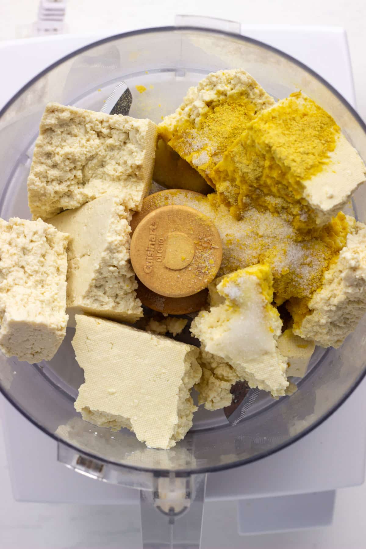 Pieces of tofu, nutritional yeast, salt and lemon juice in the bowl of a food processor before blending into Tofu Cream Cheese.