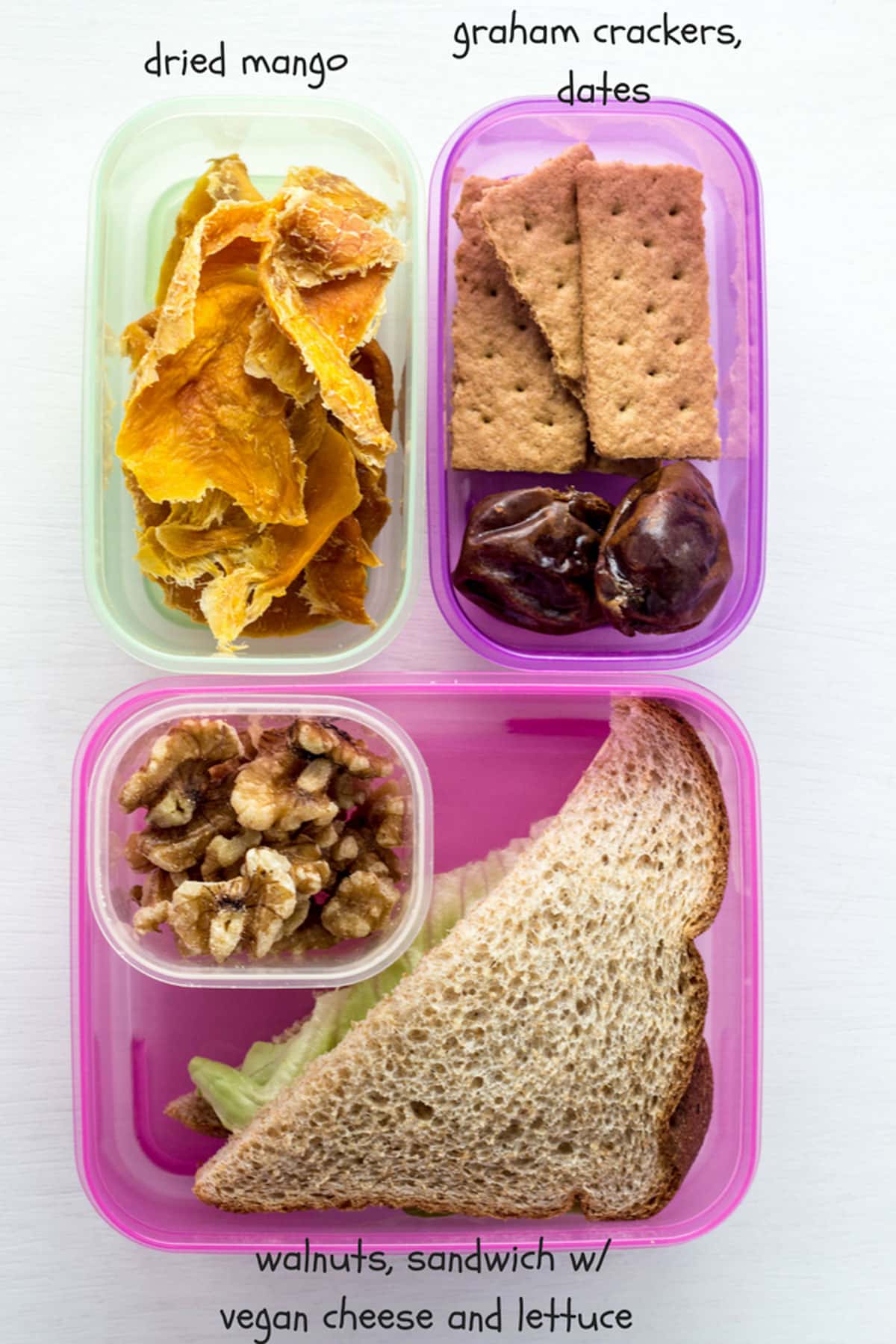 A small green container with dried mango, a small purple container with graham crackers and dates and a larger pink container with half of a sandwich and walnuts.