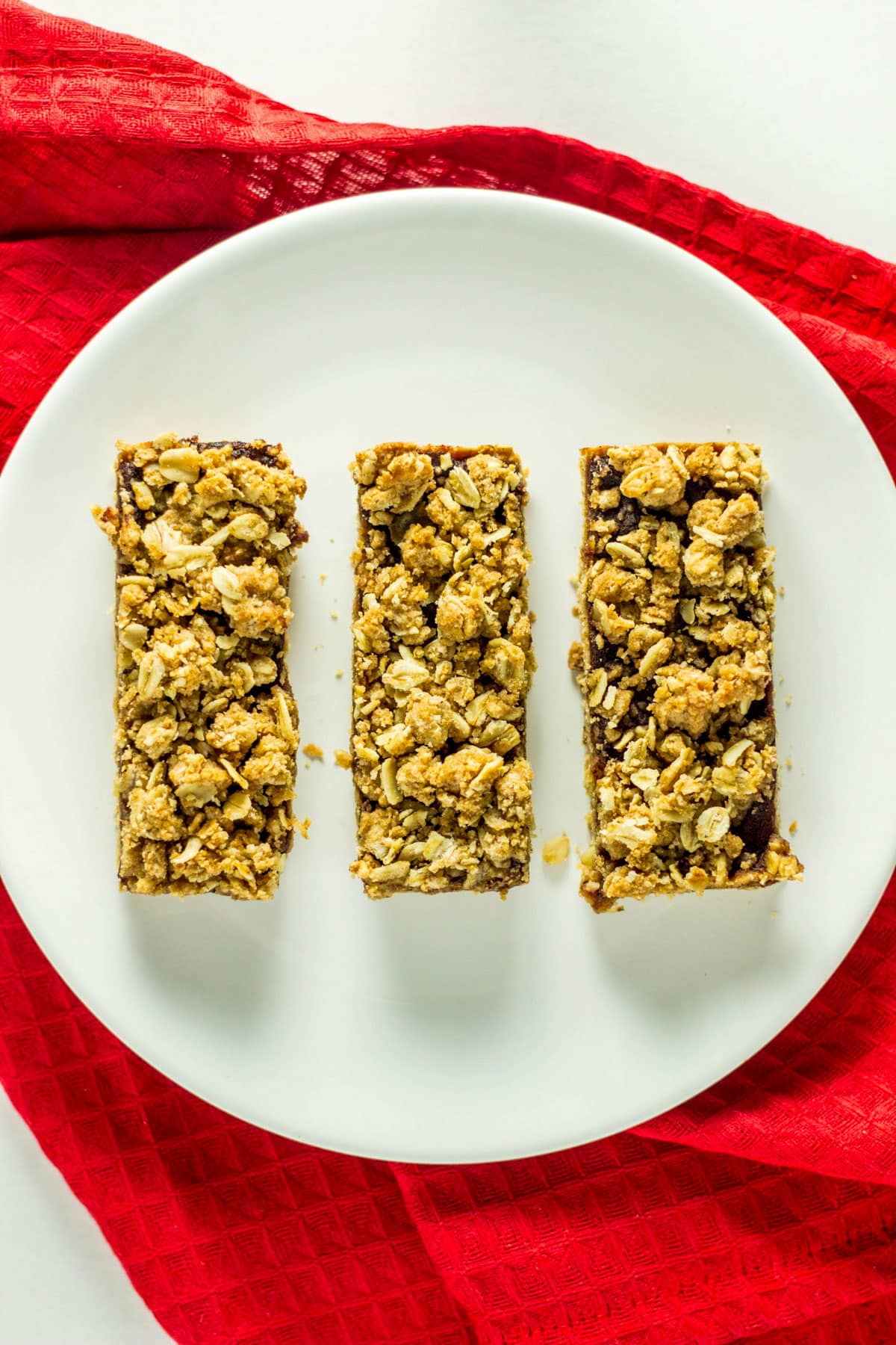 Three Vegan Apple Butter Oatmeal Bars lined up on a small white plate. The plate is sitting on a textured red towel.