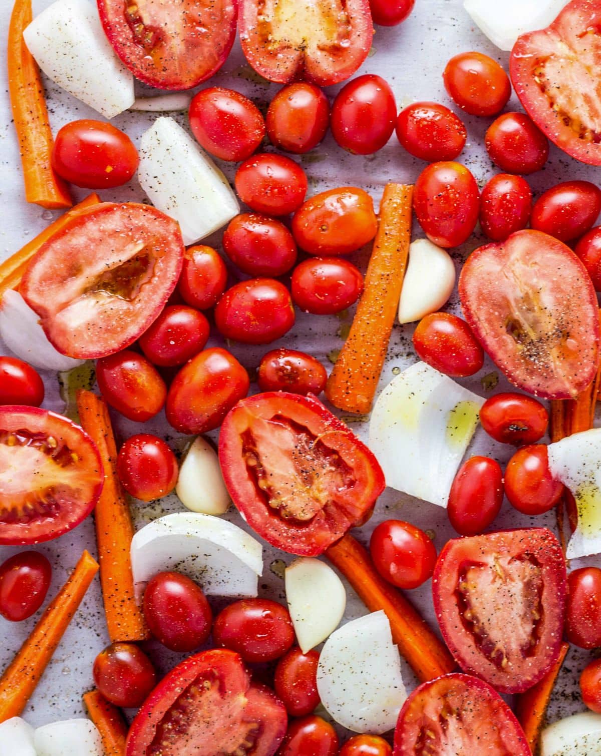 Raw Tomato halves, grape tomatoes, onions and carrots with oil, salt and pepper on a baking sheet.