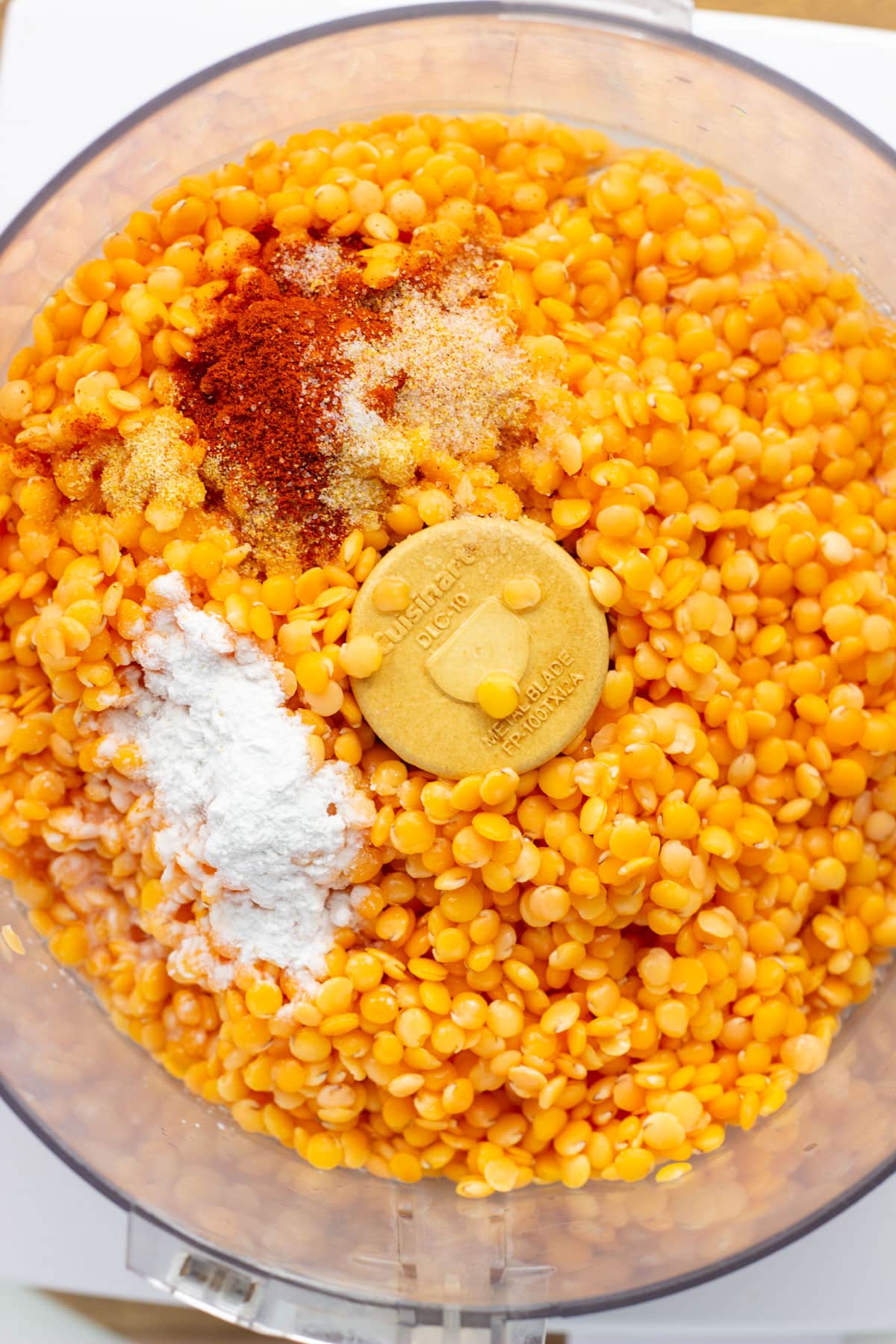 Overhead view of red lentils, spices, baking powder oil and water in the bowl of food processor.