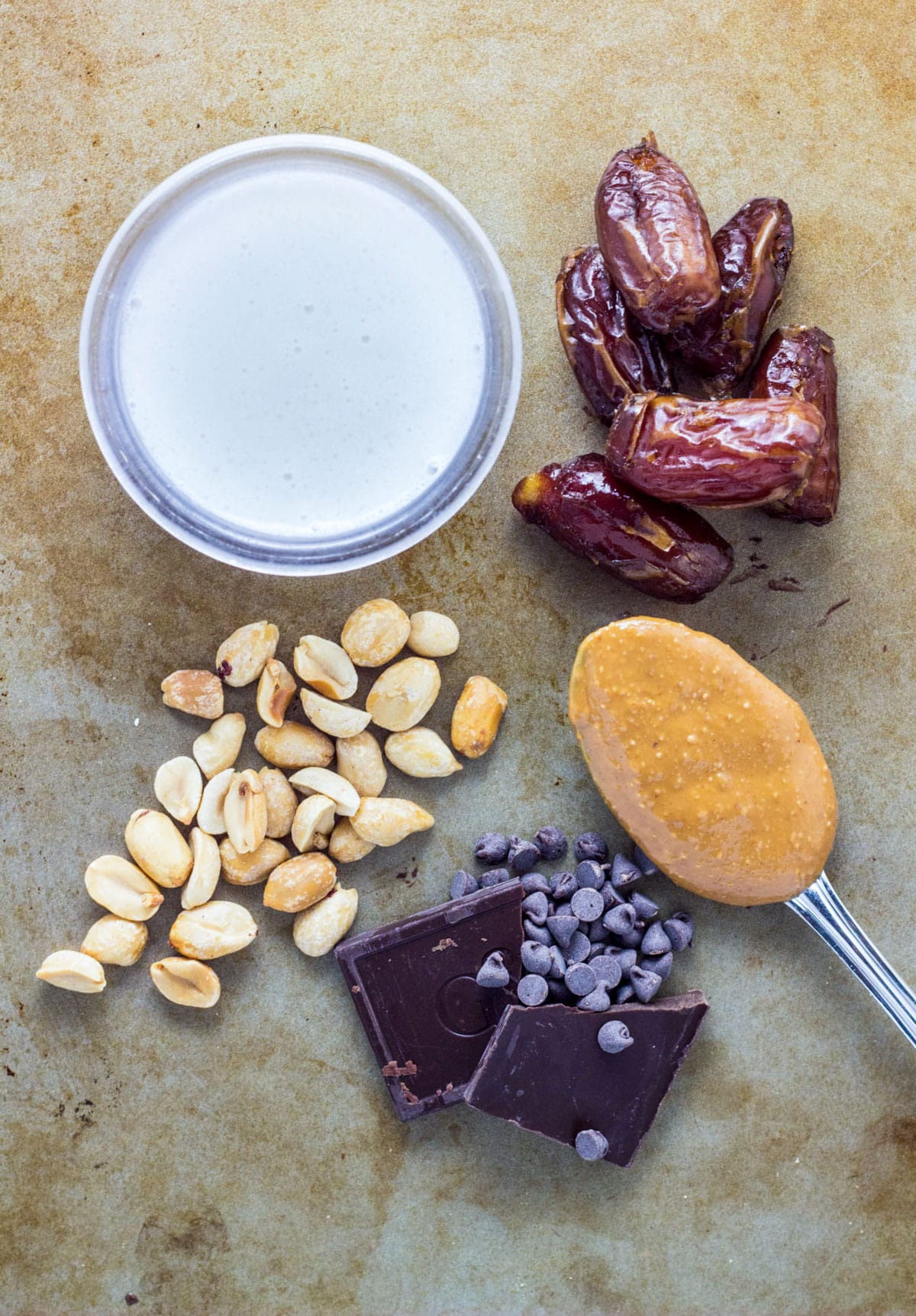 Overhead view of chocolate, peanut butter on a spoon, dates. coconut milk in a small bowl and roasted peanuts on a sheet pan.