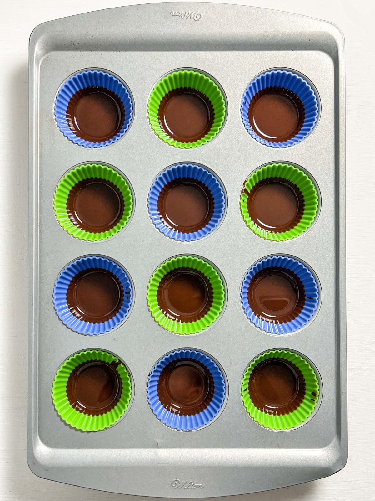 Overhead view of a muffin pan with colorful silicone liners with a layer of melted chocolate.