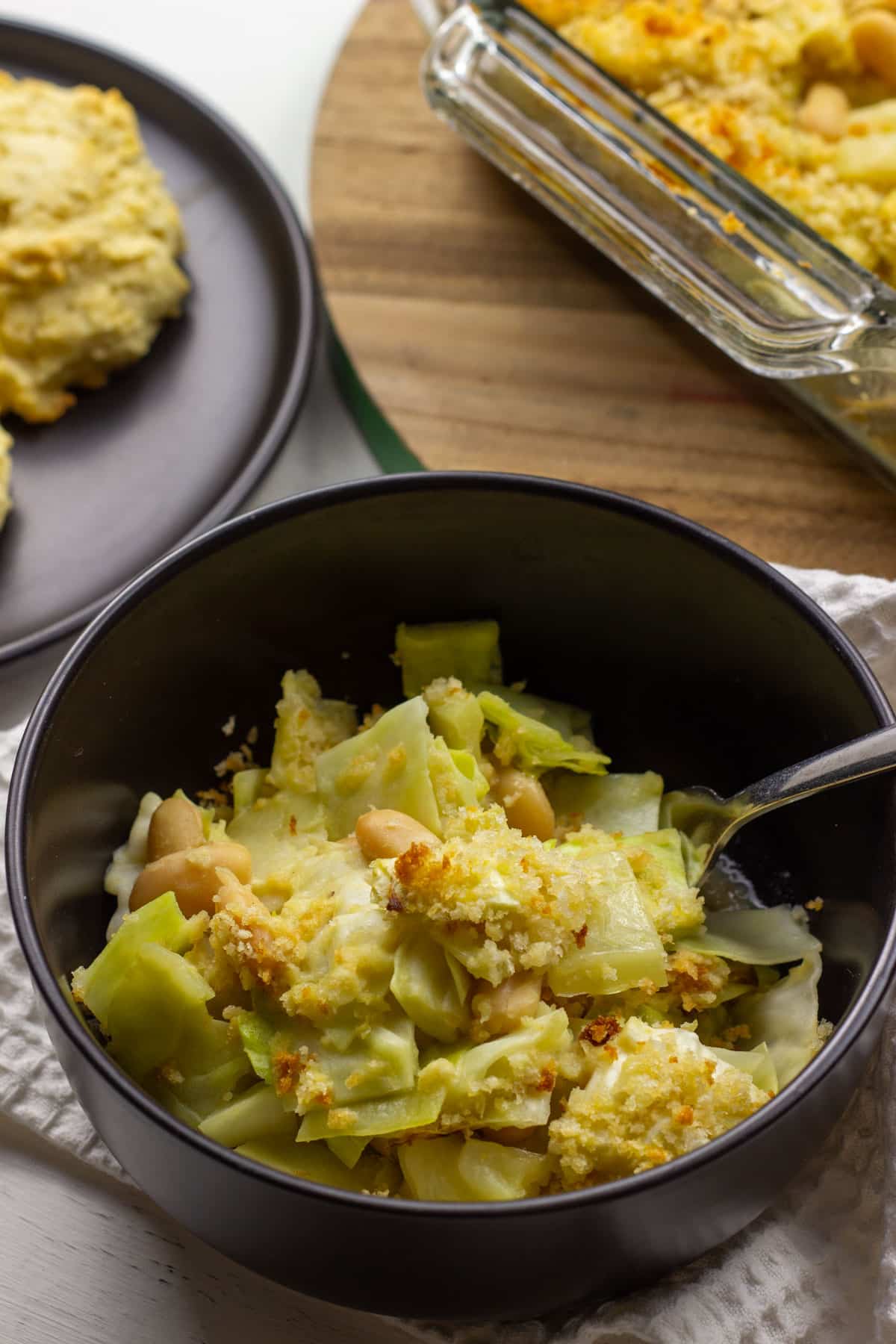 Close up view of a bowl of Vegan Scalloped Cabbage and Beans. There is a casserole dish partially visible in the upper right and a plate with biscuits to the left.