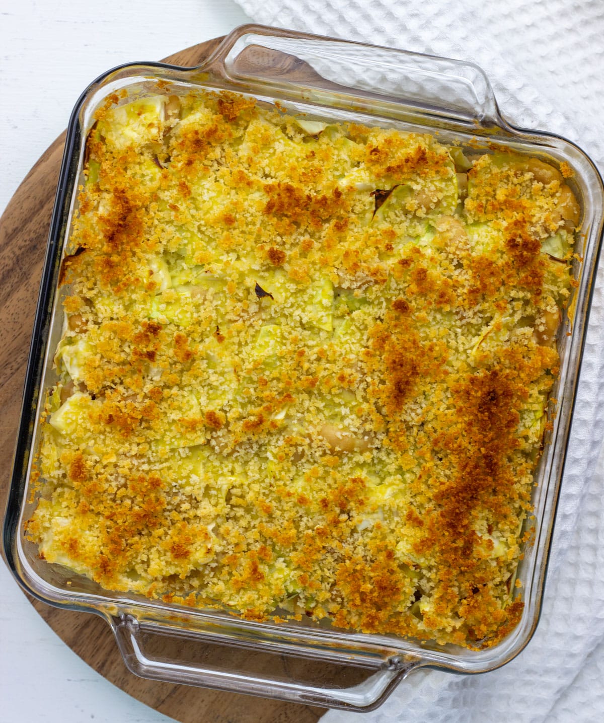 Overhead view of Vegan Scalloped Cabbage and Beans in a baking dish. The dish sits on a round wooden board with a white textured towel to the right.