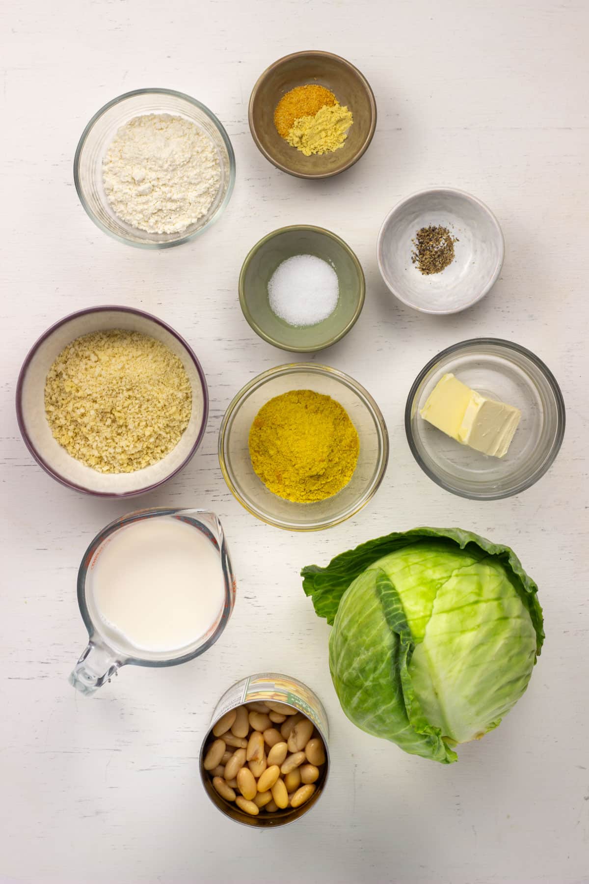 Ingredients to make Vegan Scalloped Cabbage and Beans. There is green cabbage, cannellini beans, non dairy milk, nutritional yeast, non dairy butter, bread crumbs, salt, pepper, all purpose flour, garlic powder and ground mustard.