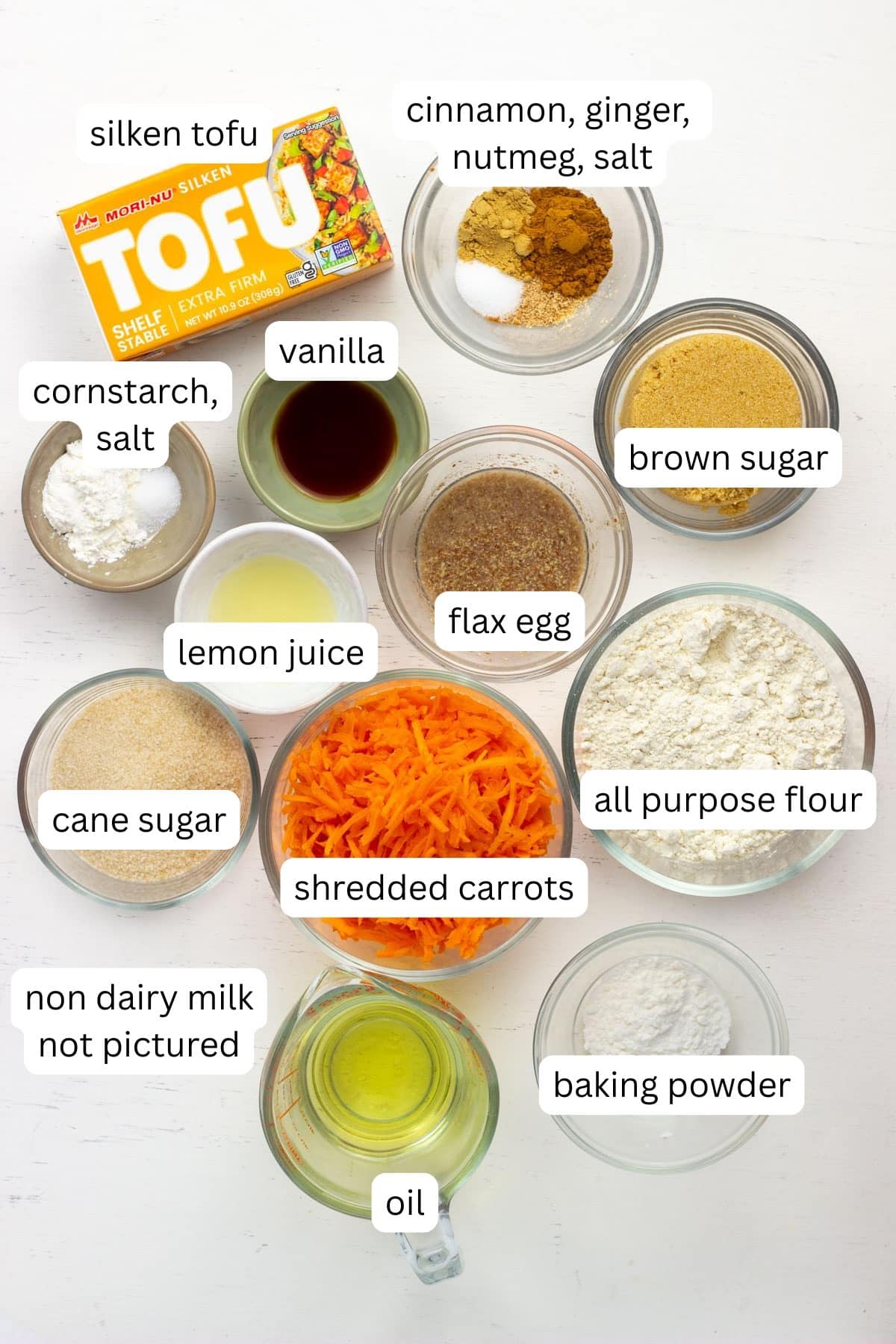 Ingredients to make Vegan Carrot Cake Cheesecake Bars. There are labeled bowls of shredded carrots, flour, sugar, brown sugar, baking powder, oil, lemon juice, vanilla extract, flax egg, spices and a box of shelf stable tofu.