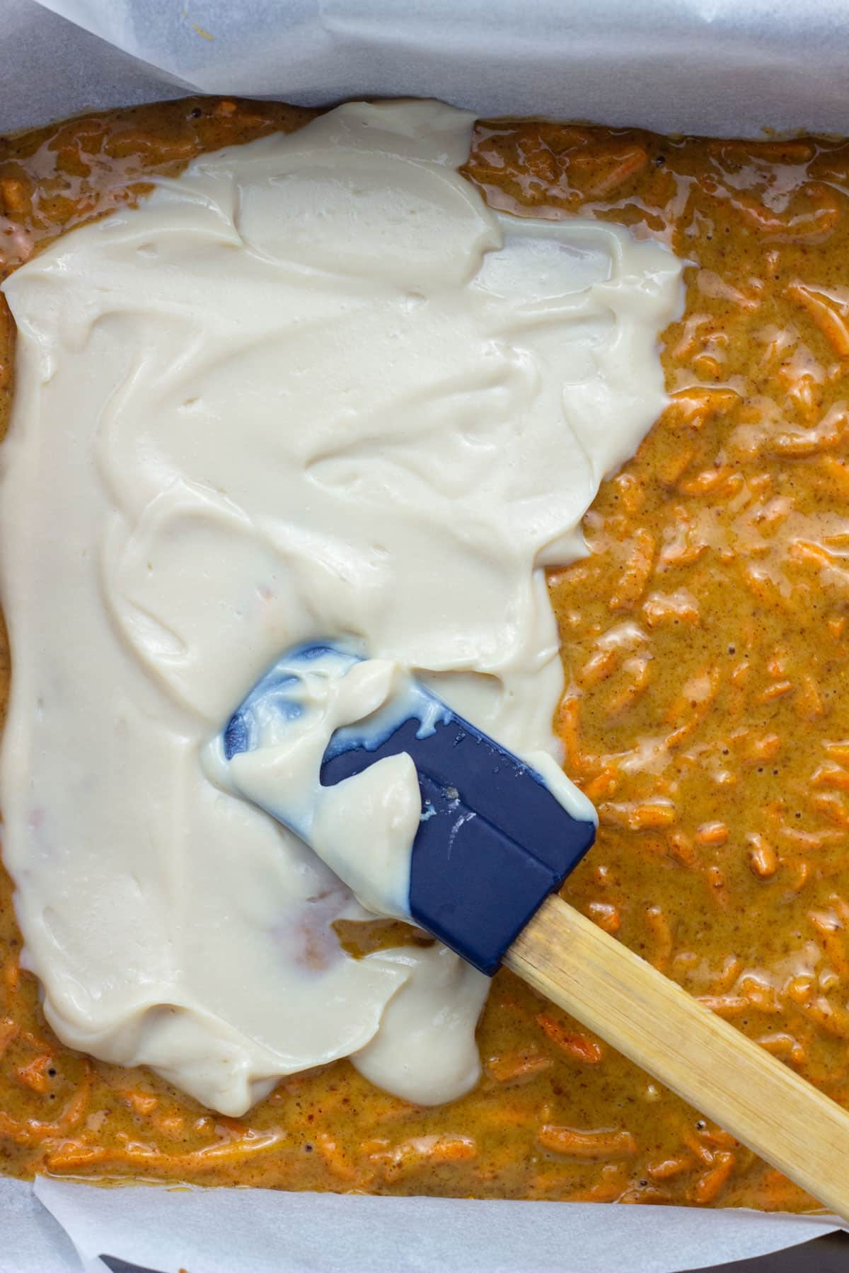Close up of carrot cake batter in a baking pan. There is cheesecake filling being spread over the top of batter with a small spatula.