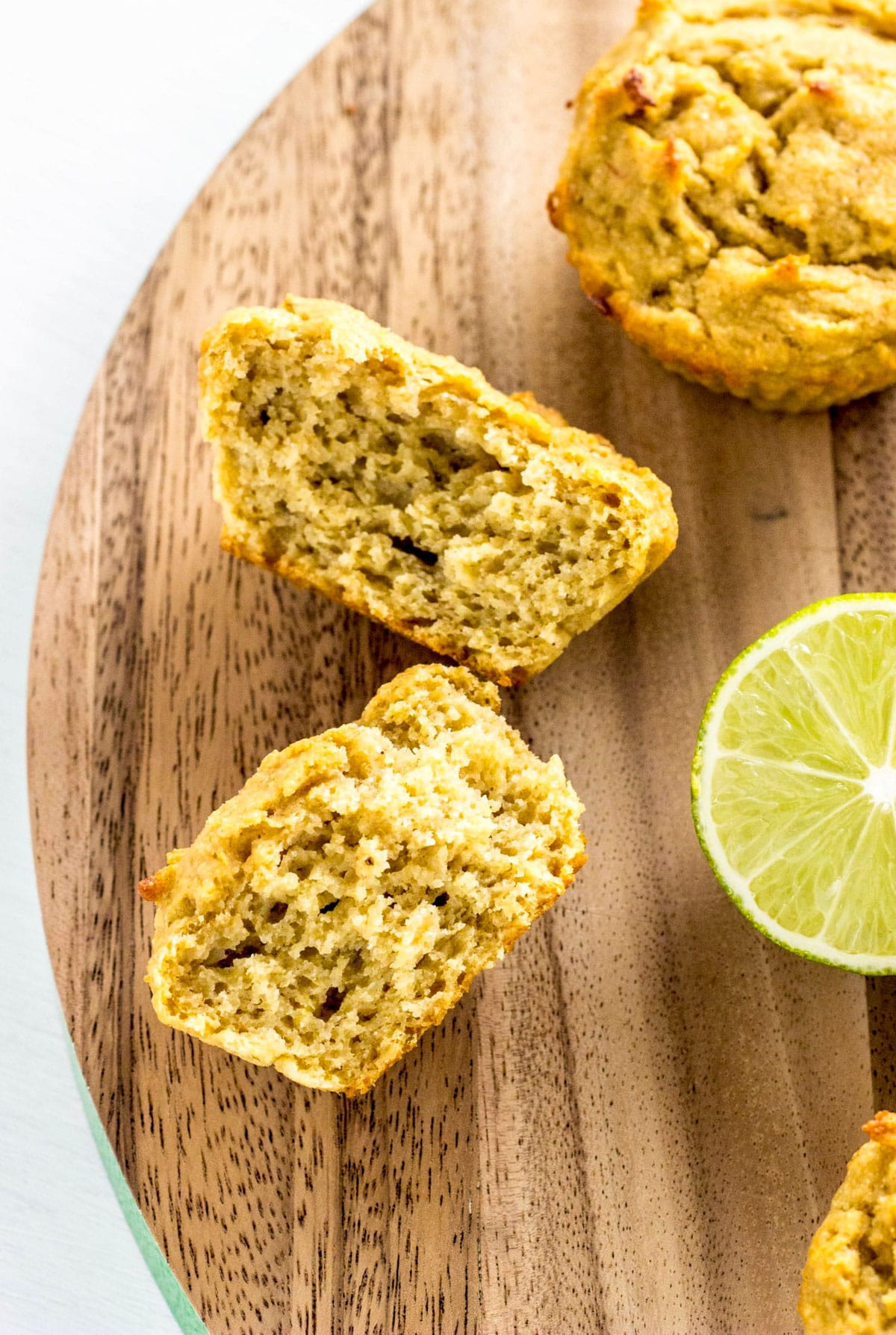A Vegan Lime Avocado Muffin cut in half to show interior texture. Other muffins and a lime are partially visible in the frame.
