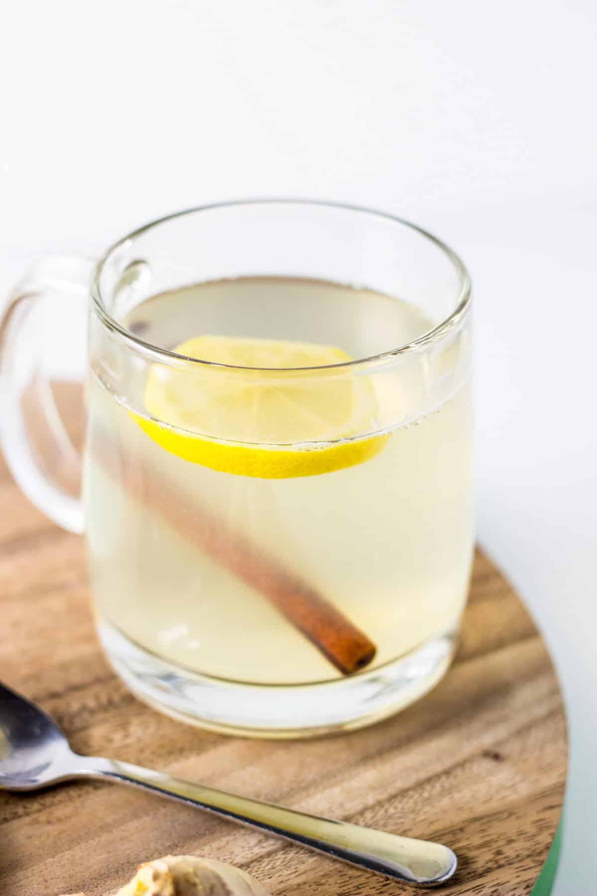 A clear mug of soothing Ginger Lemon Tea. There is a slice of lemon and a cinnamon stick in the mug.