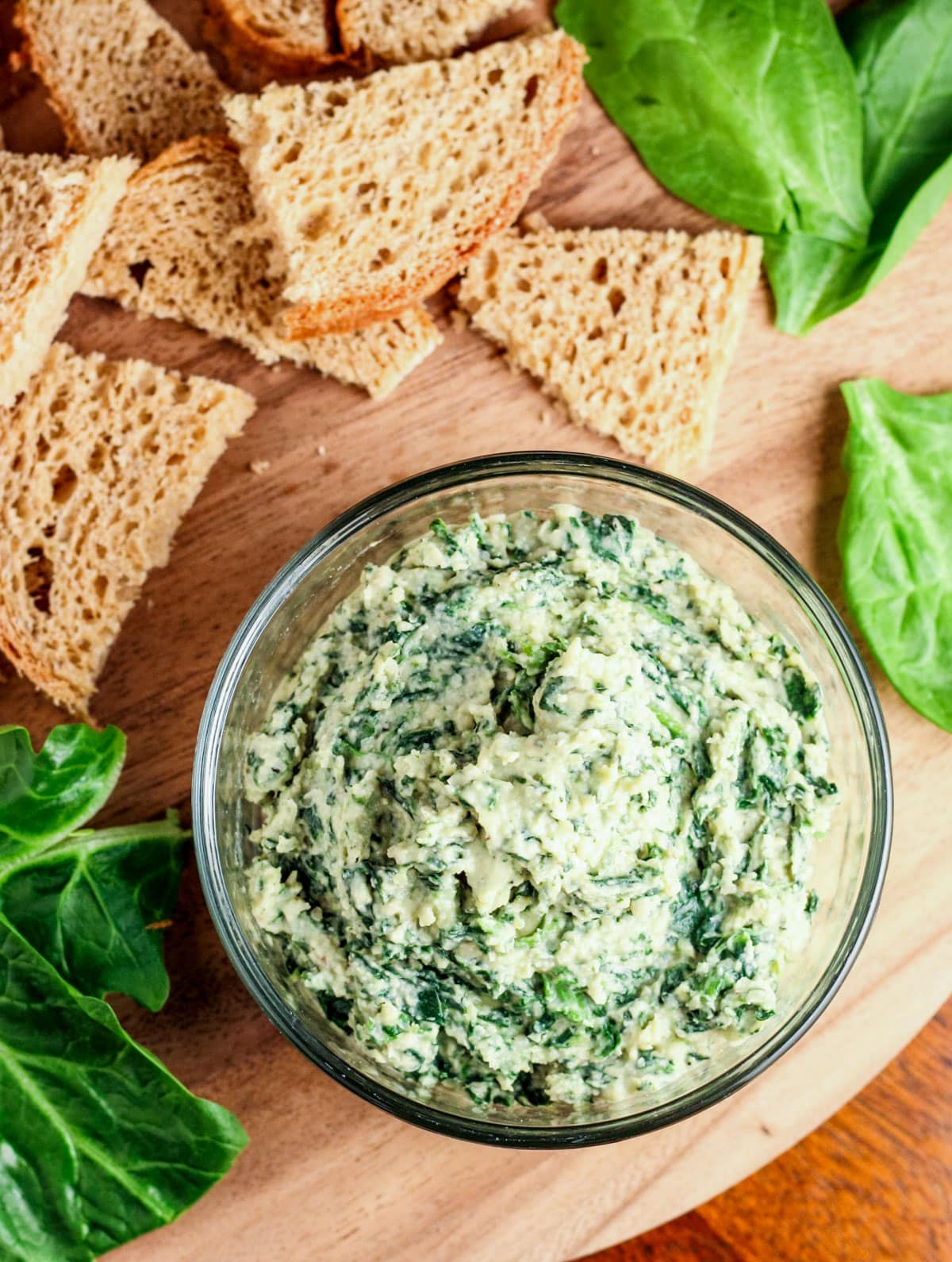Top view of a bowl of Vegan Spinach White Bean Dip with pieces of bread and spinach around.