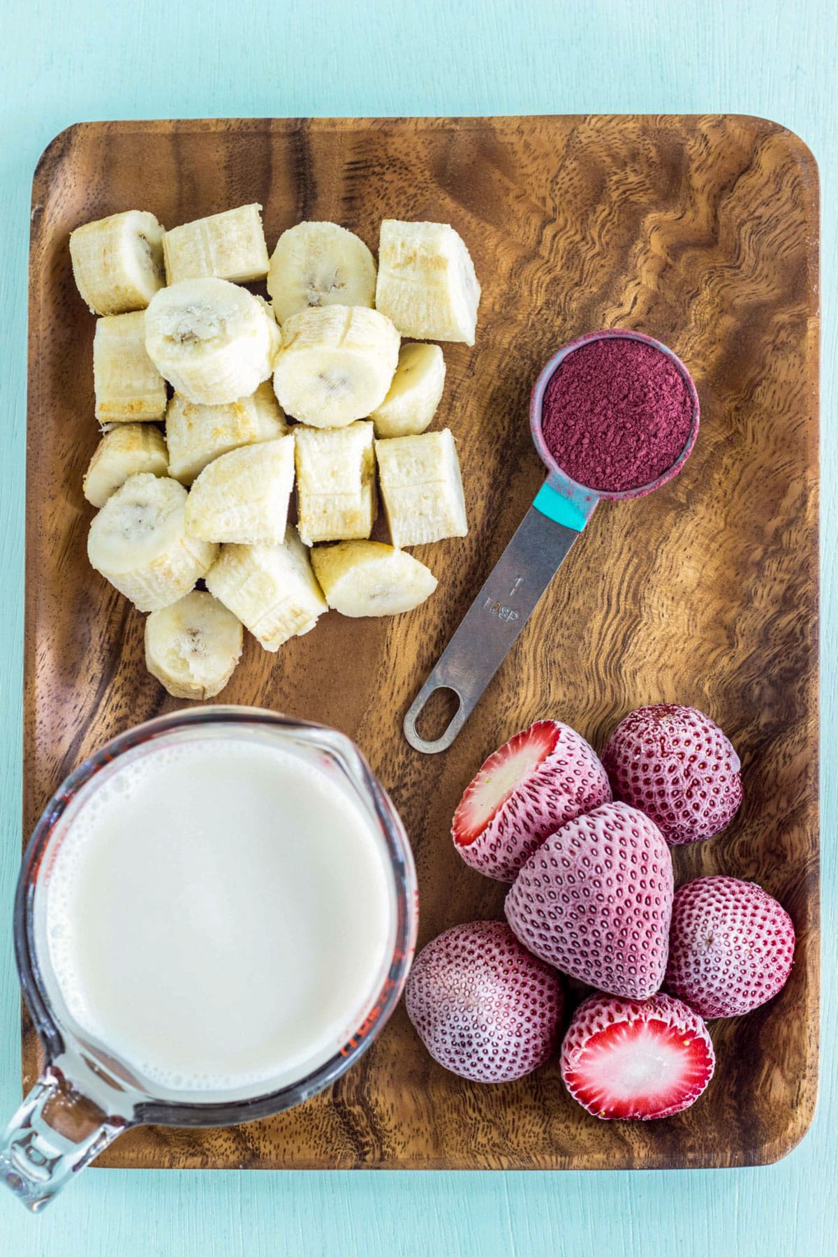 Frozen strawberries, frozen banana slices, beet powder and non dairy milk on a wooden board.