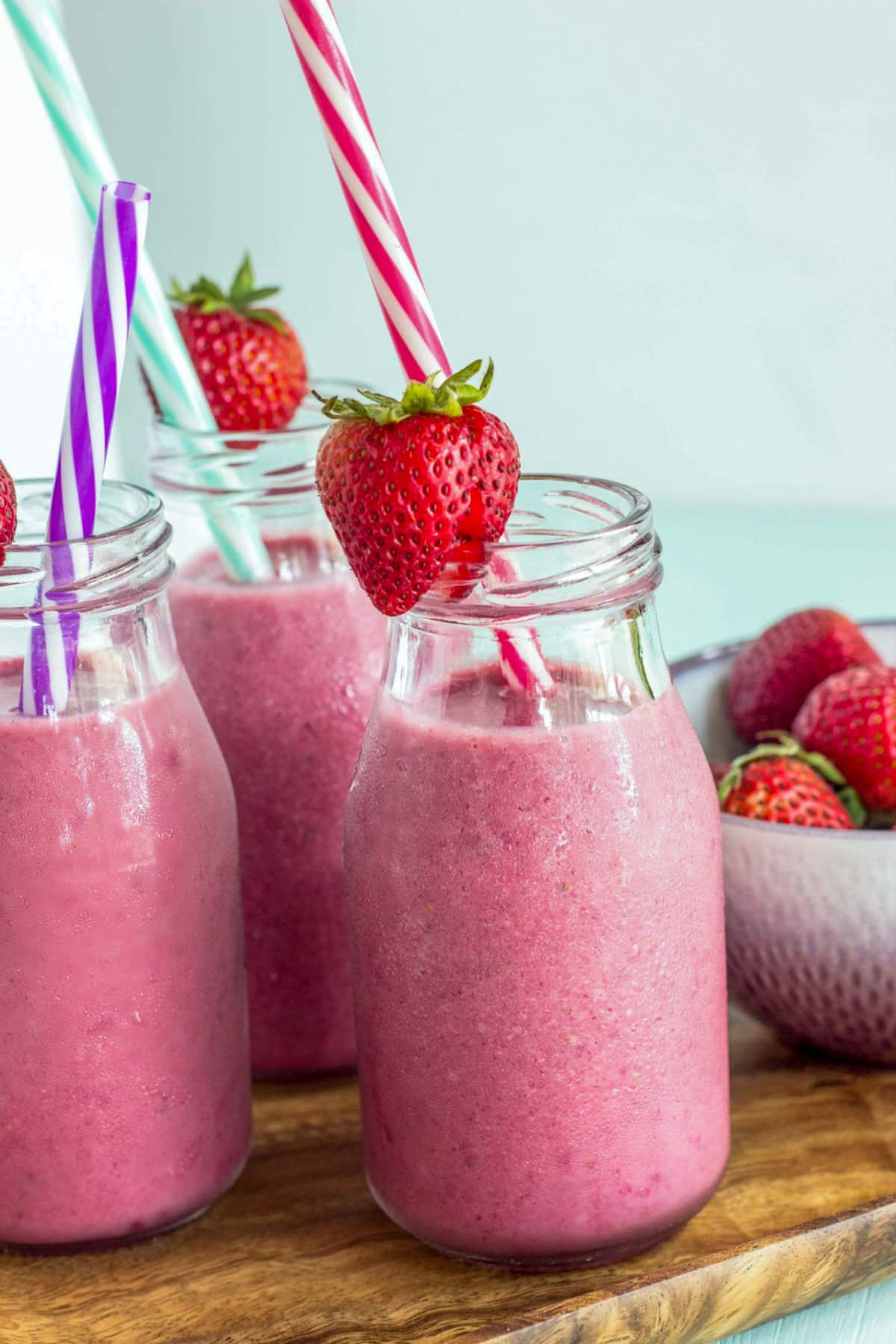 Small bottles of Strawberry Banana Beet Smoothie sitting on a wooden board. The bottles have straws and a strawberry on the rim.