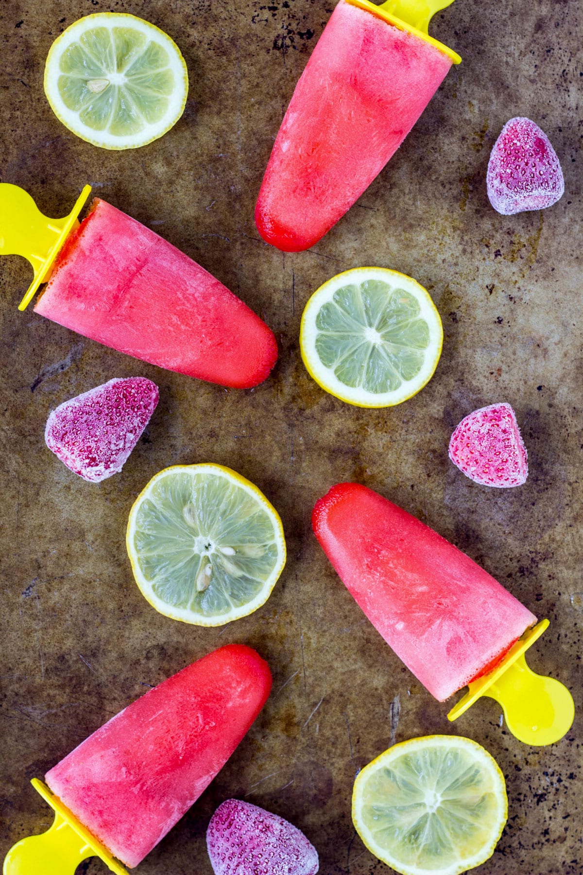 Four Strawberry Lemonade Popsicles on a sheet pan with lemon slices and frozen strawberries.